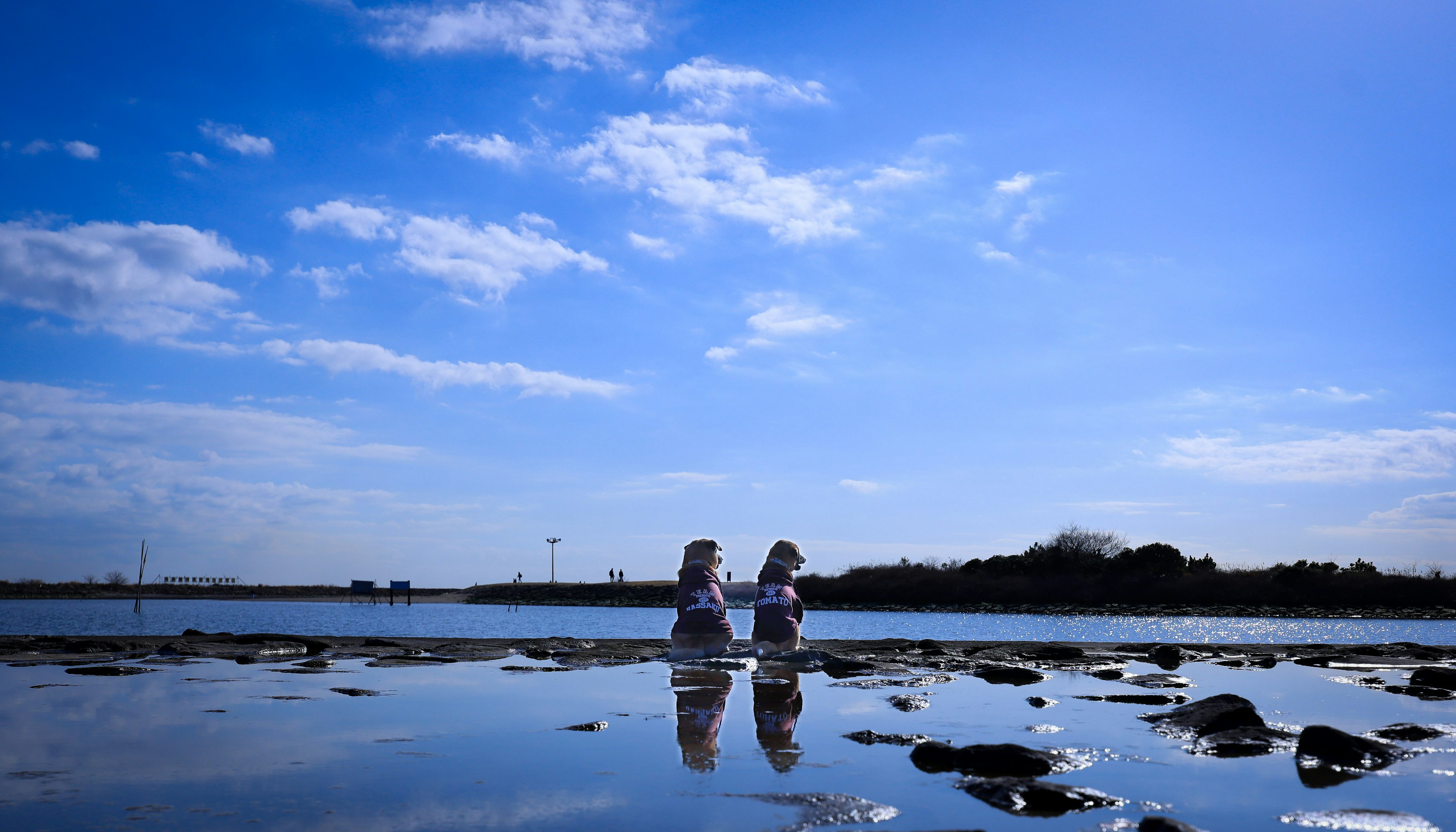 Dos mujeres de pie junto al agua bajo un cielo azul reflejándose en la superficie