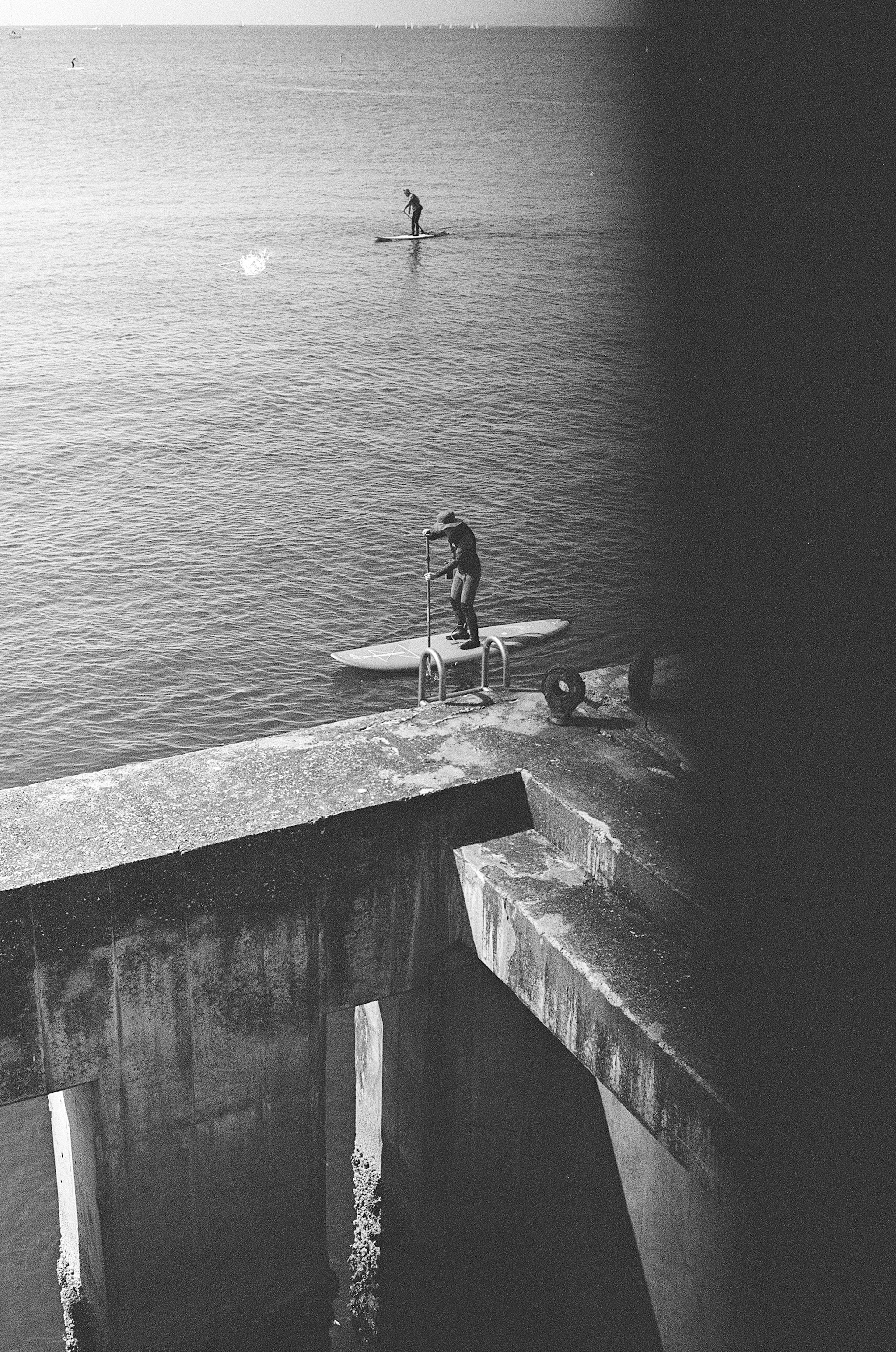 Black and white photo of a person standing on a pier with another person surfing on the water