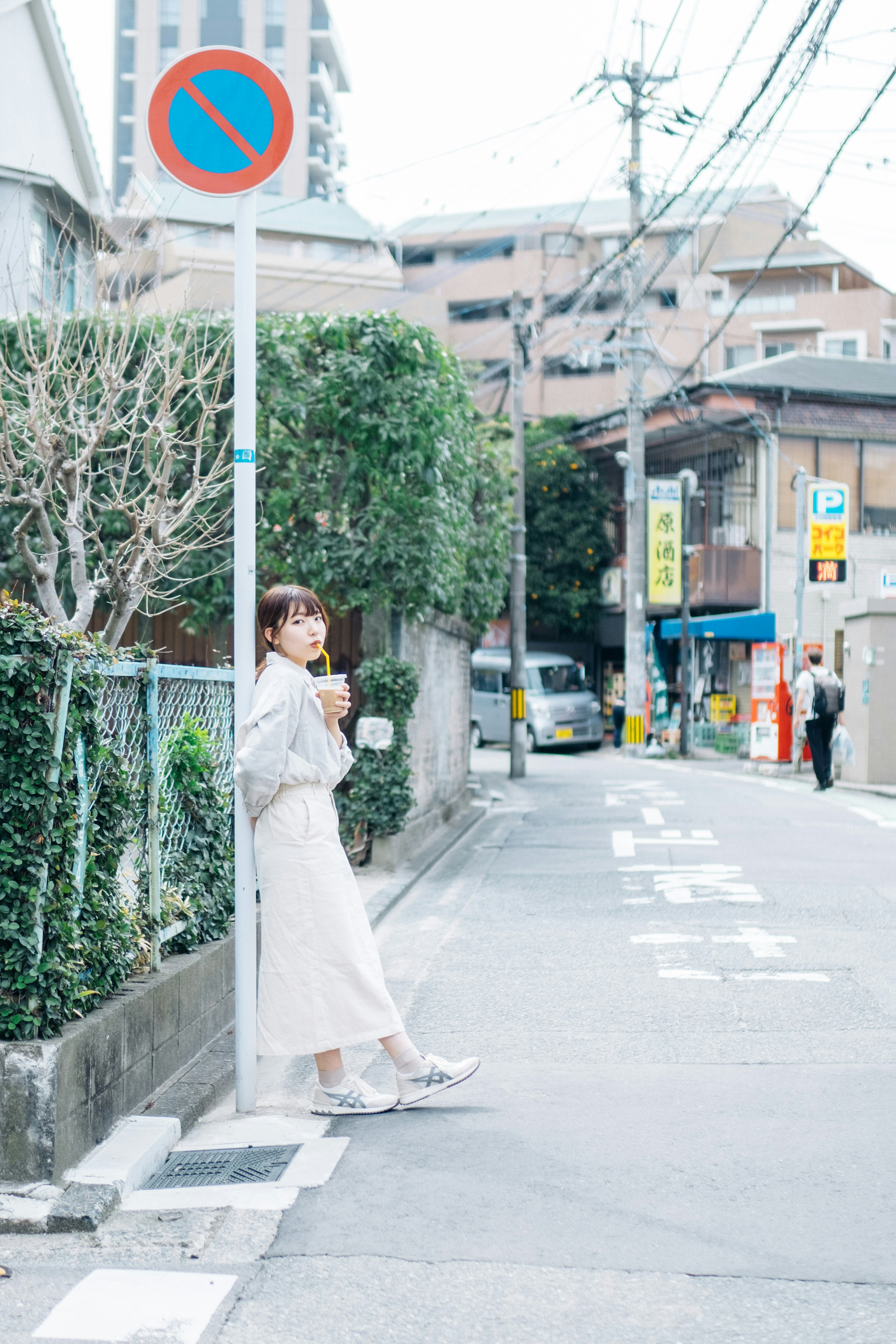 A woman standing on a street corner wearing white clothing surrounded by greenery and a no parking sign