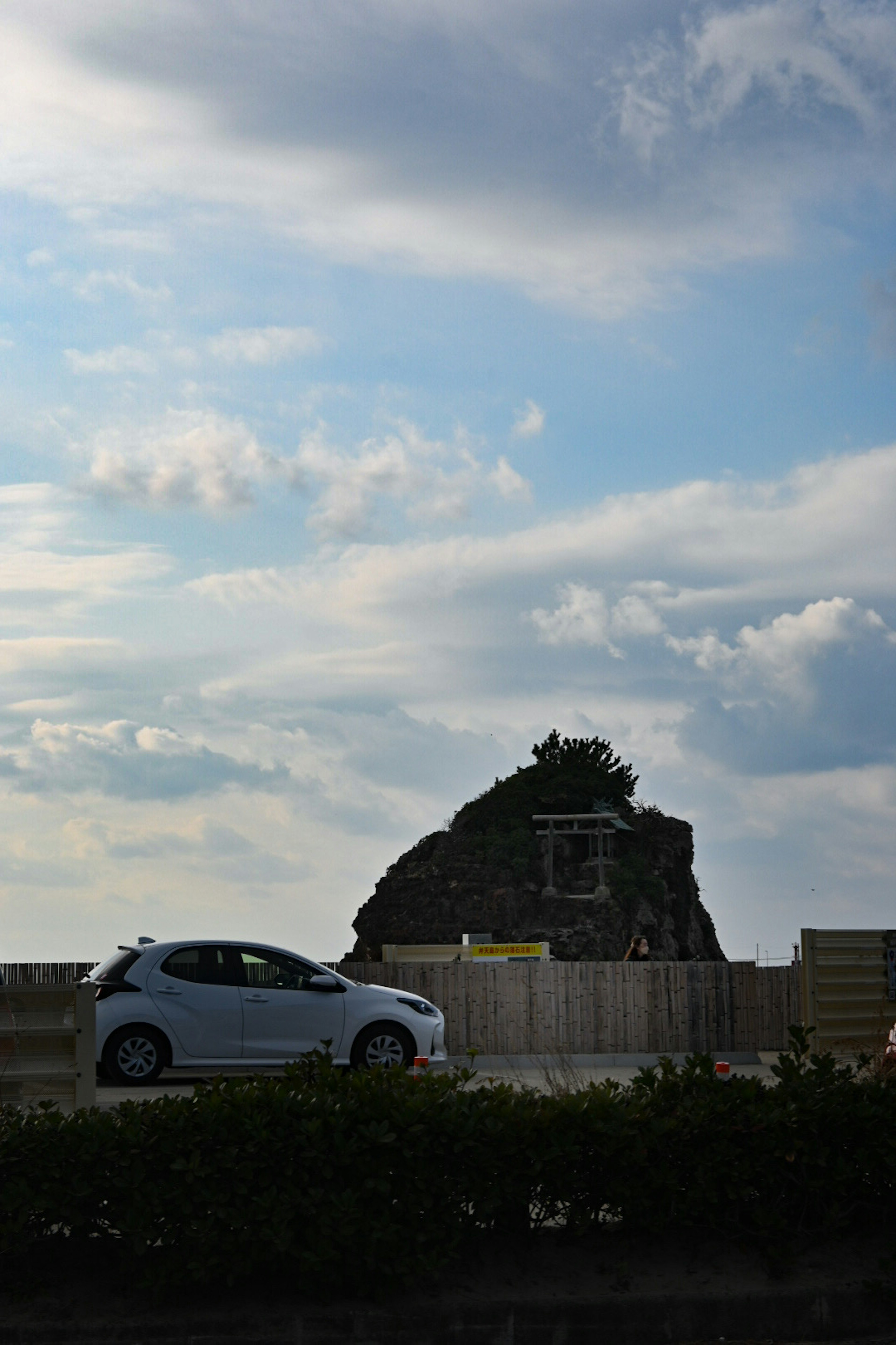 Ein weißes Auto fährt an einem Felsen mit Grün unter einem bewölkten Himmel vorbei