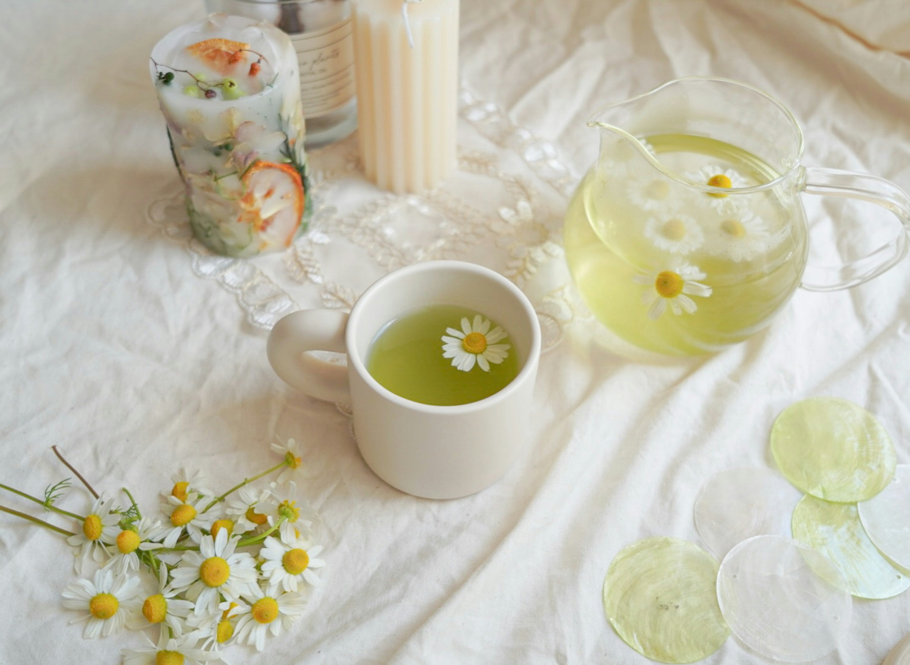 Una tazza di tè verde con un fiore di camomilla galleggiante accanto a una caraffa di tè e involtini di insalata freschi su un tessuto bianco