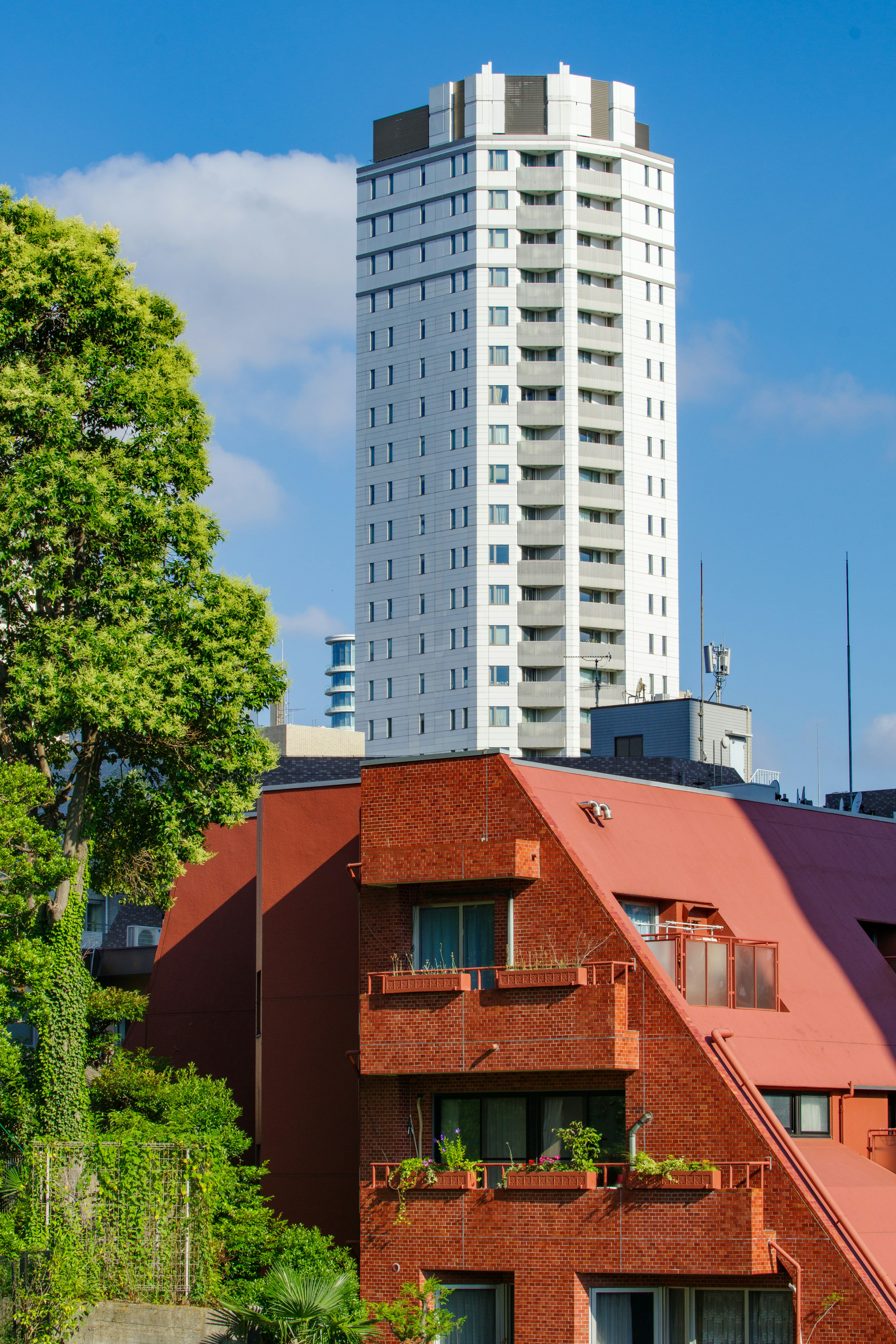 Pemandangan cerah dengan bangunan merah di samping gedung pencakar langit putih dikelilingi oleh pepohonan hijau dan langit biru