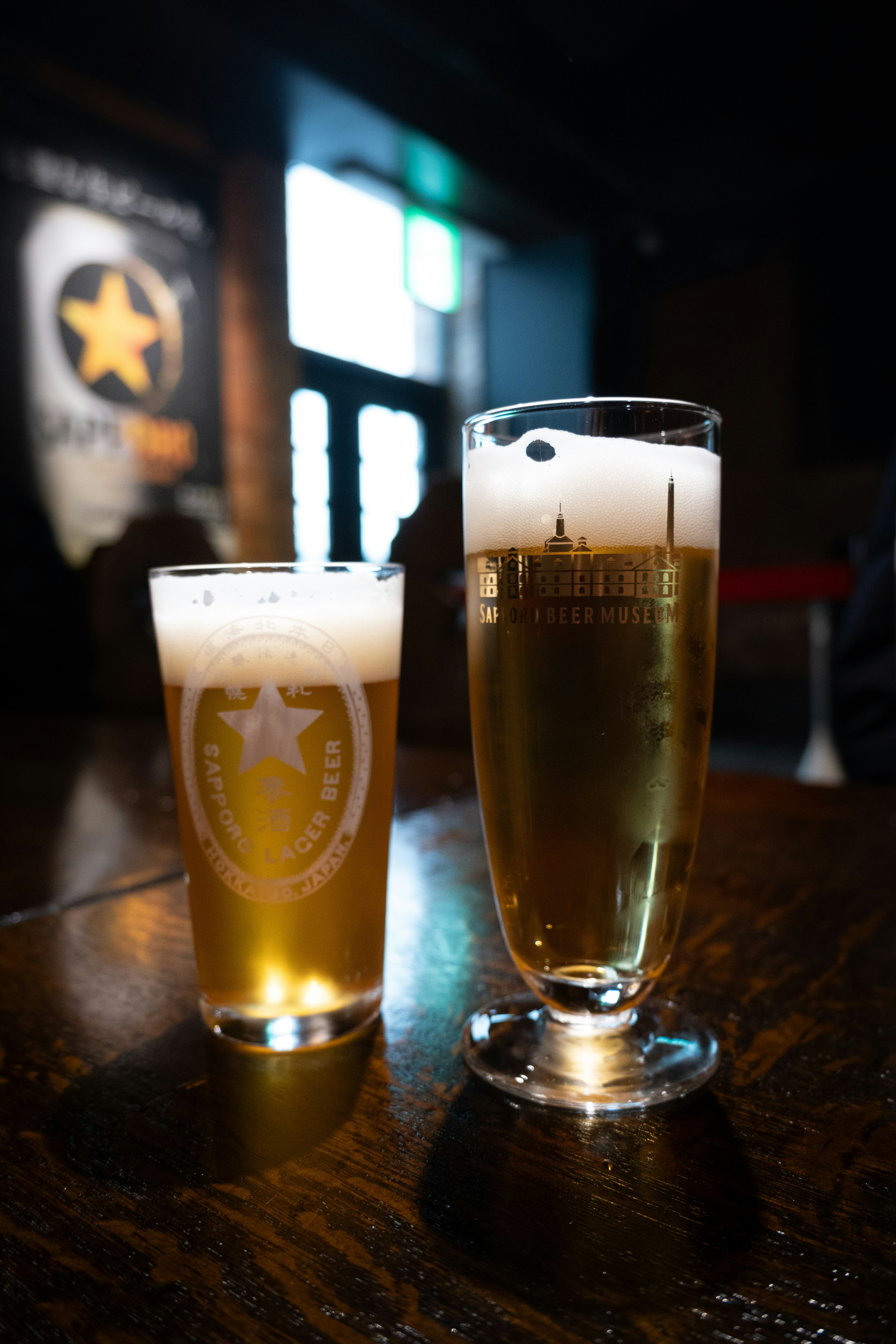 Una foto que muestra dos vasos de cerveza sobre una mesa el vaso de la izquierda tiene espuma blanca y es más pequeño el vaso de la derecha es más grande con cerveza dorada