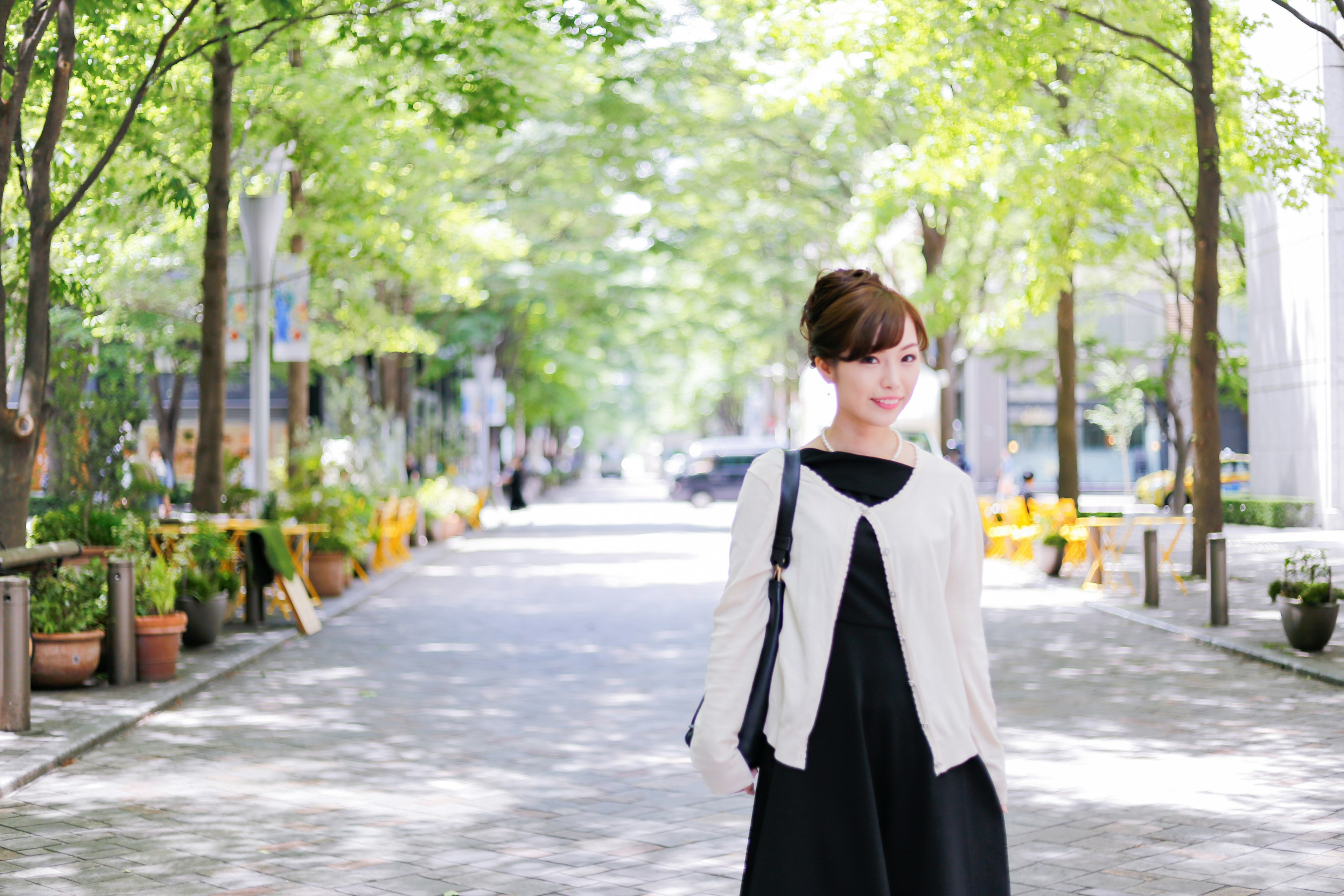 A woman standing on a green-lined street wearing a black dress and white cardigan