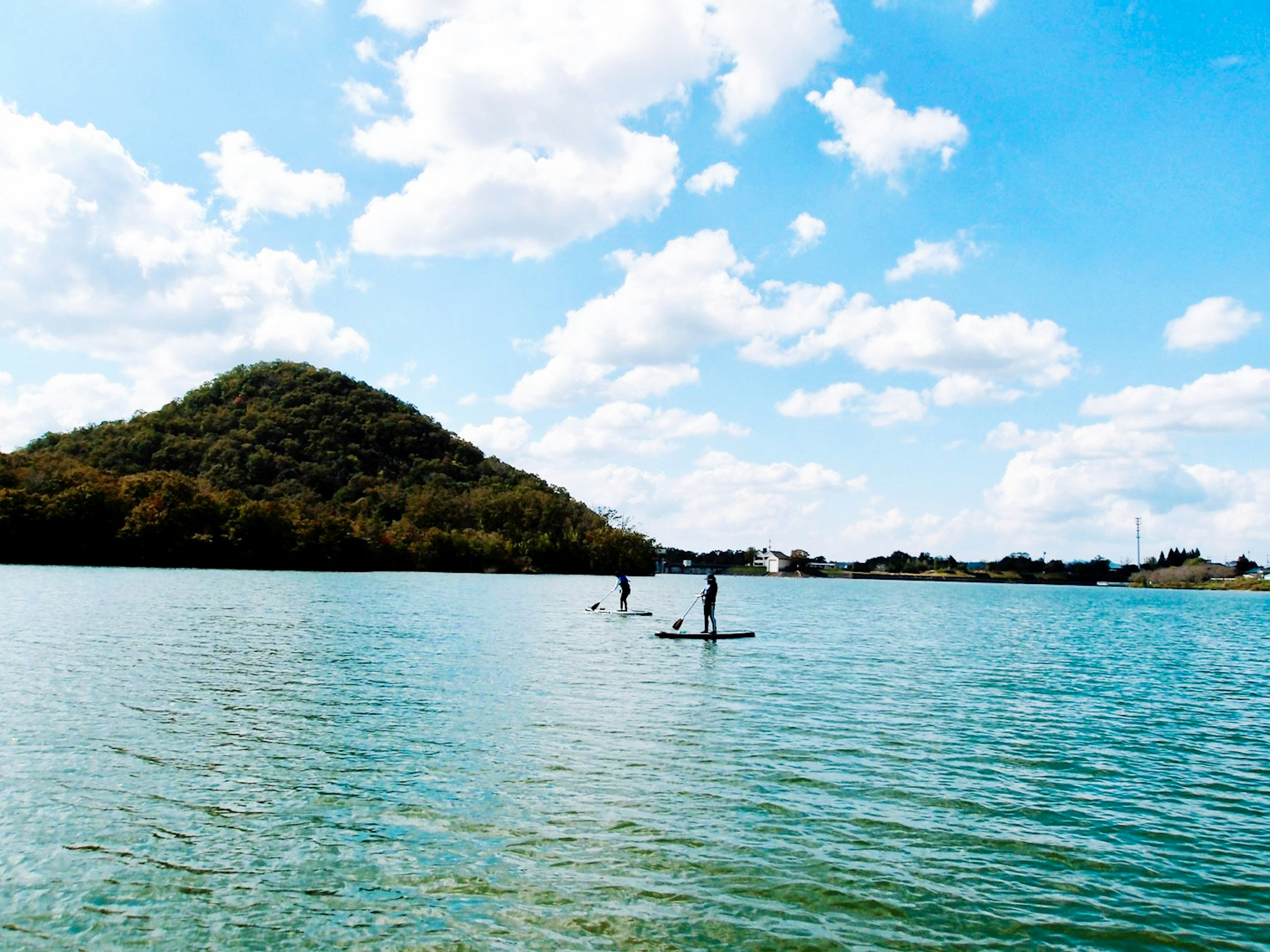 Dos personas en paddle surf en una superficie de agua tranquila bajo un cielo azul con nubes