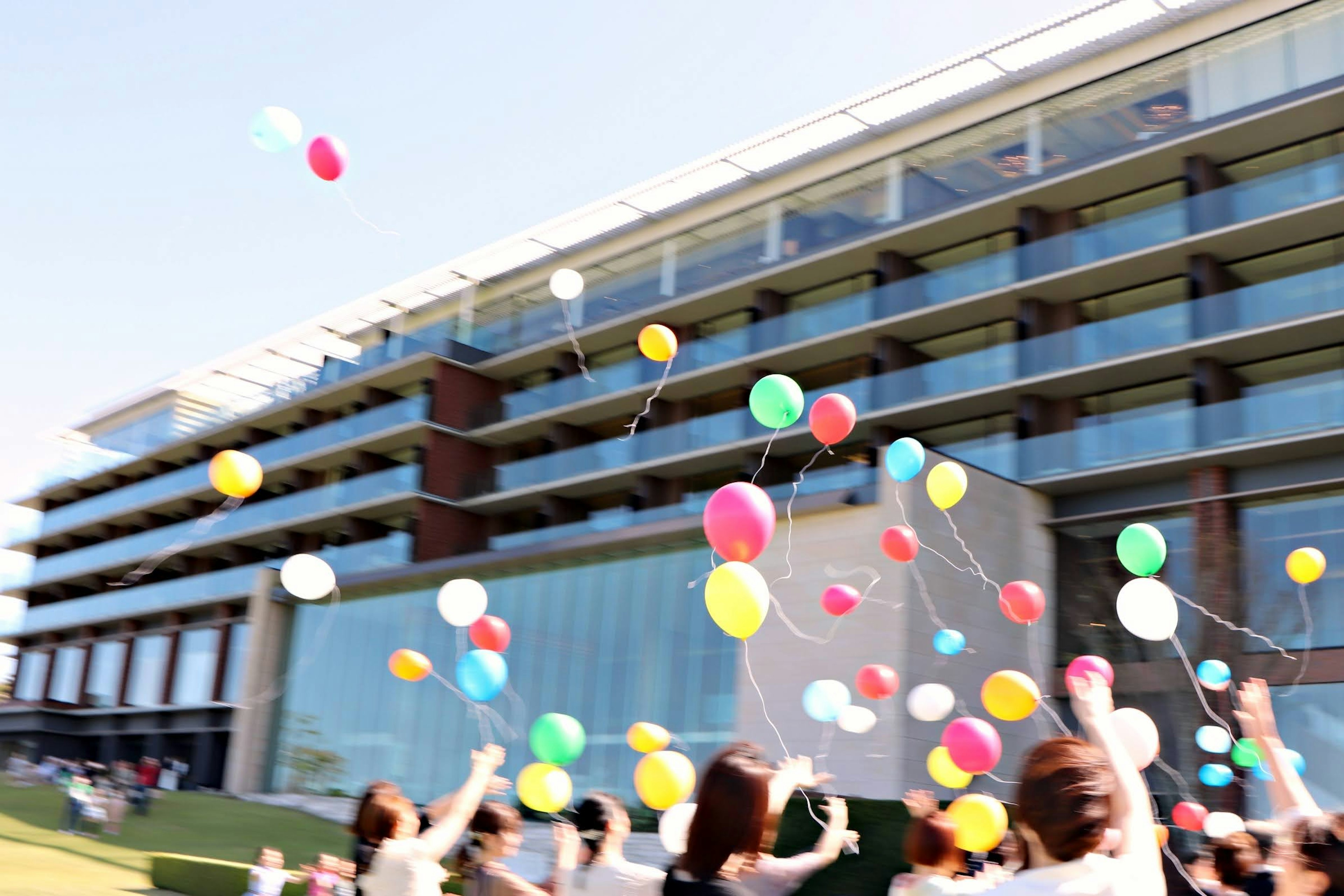 人々がカラフルな風船を空に放つイベントの様子 モダンな建物が背景にある