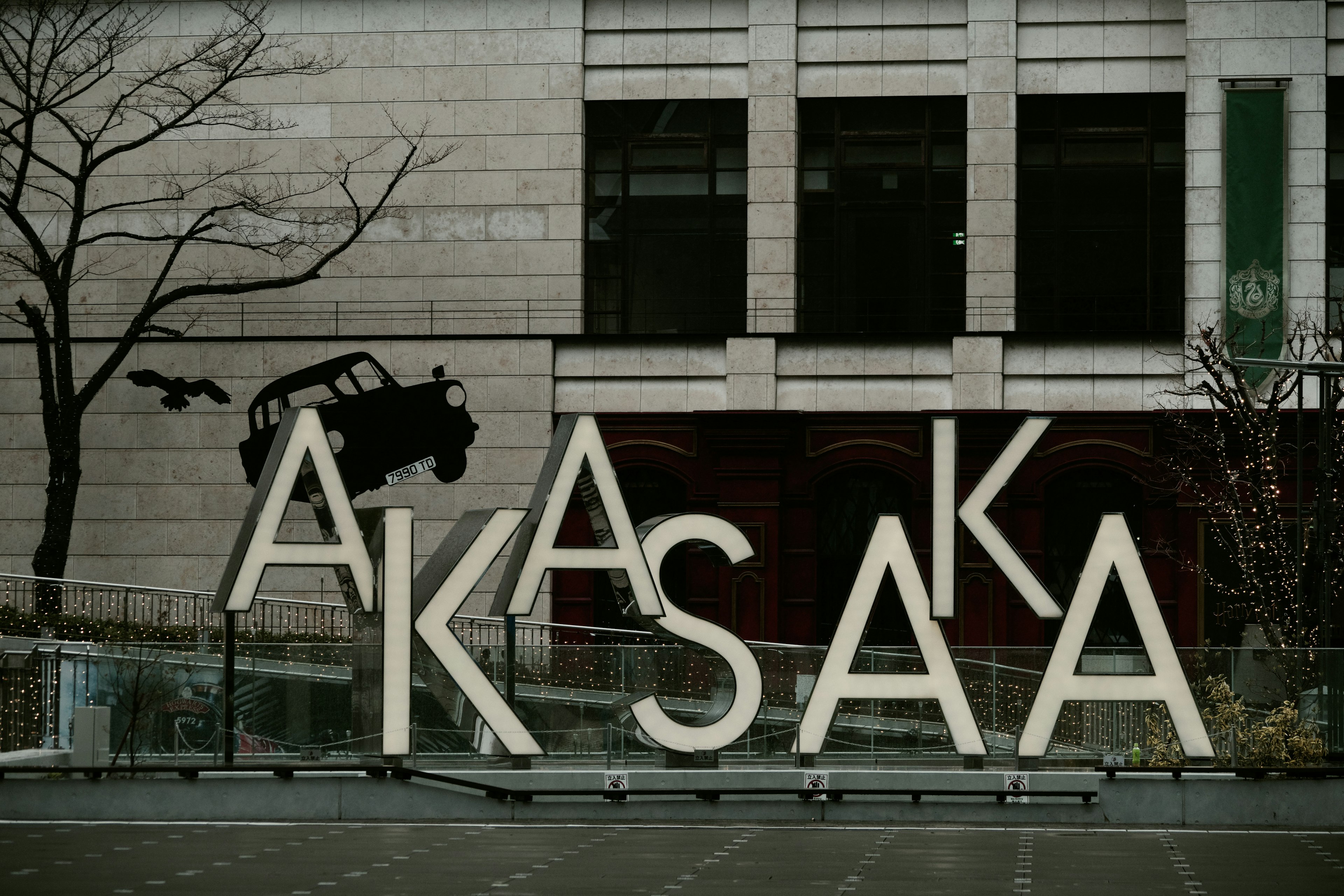 大きな文字で「AKASAKA」と書かれたアートインスタレーションの前に、木の枝と飛び去る車の影が見える