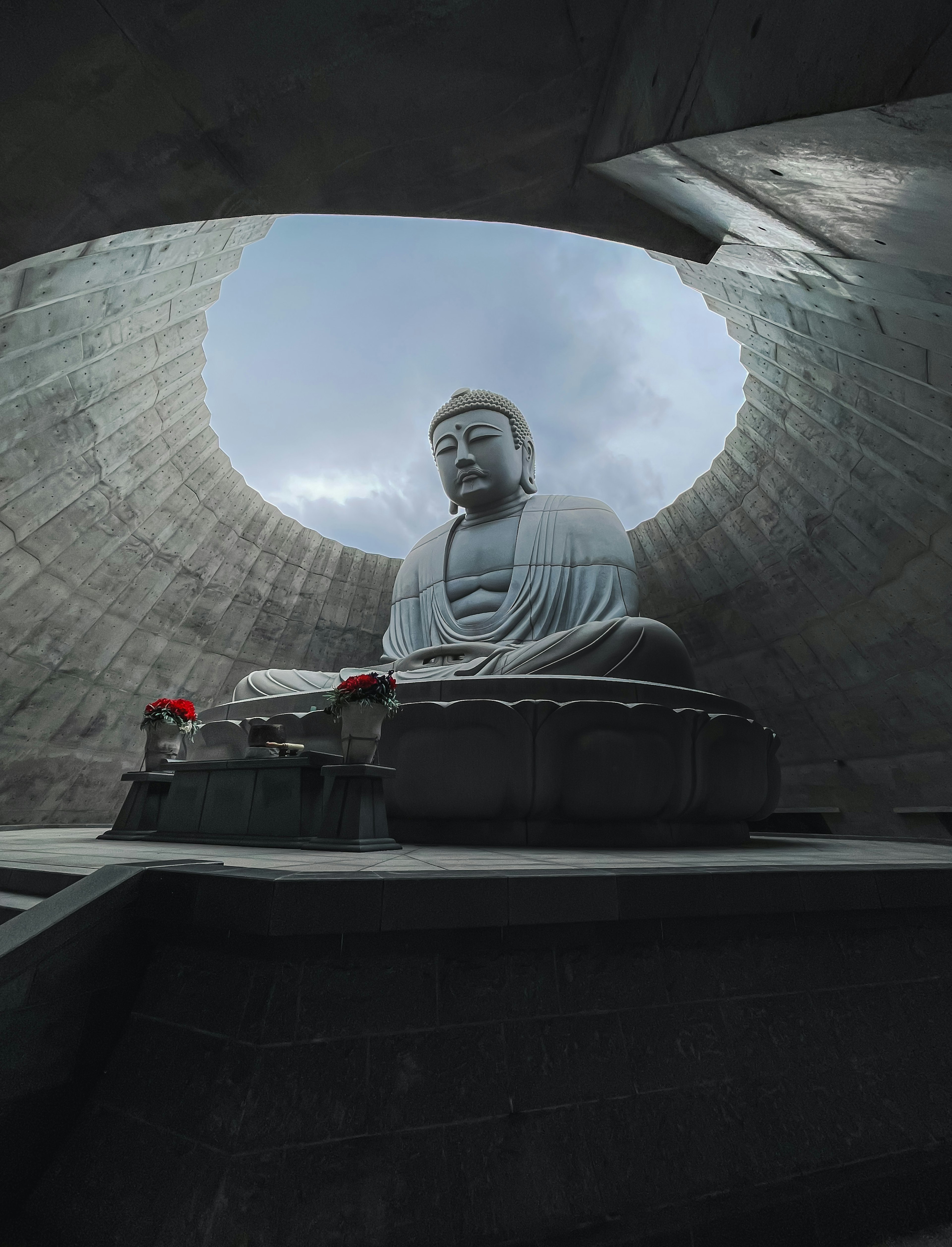 大きな仏像が立つ円形の空間の写真 雲がかかった空が見える