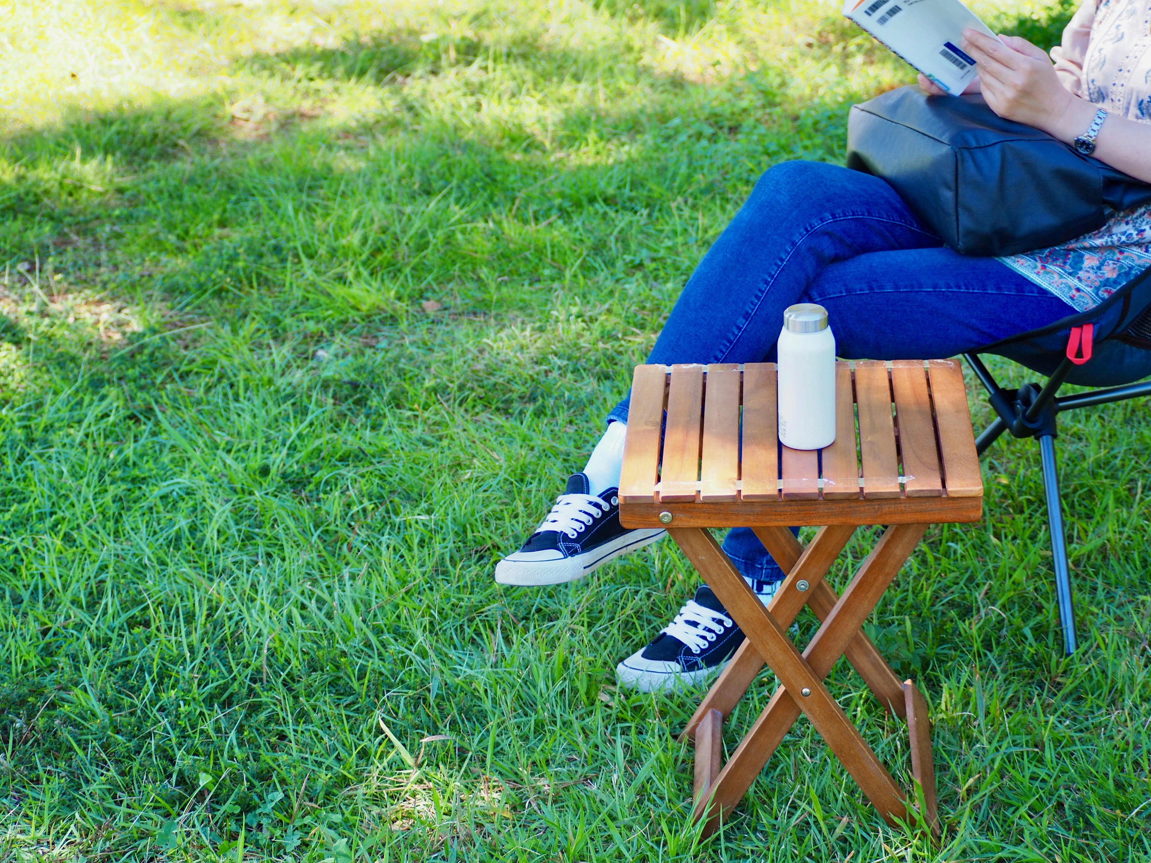 Eine Frau liest ein Magazin, sitzt auf einem Stuhl mit einem klappbaren Holztisch und einer weißen Flasche auf dem Gras