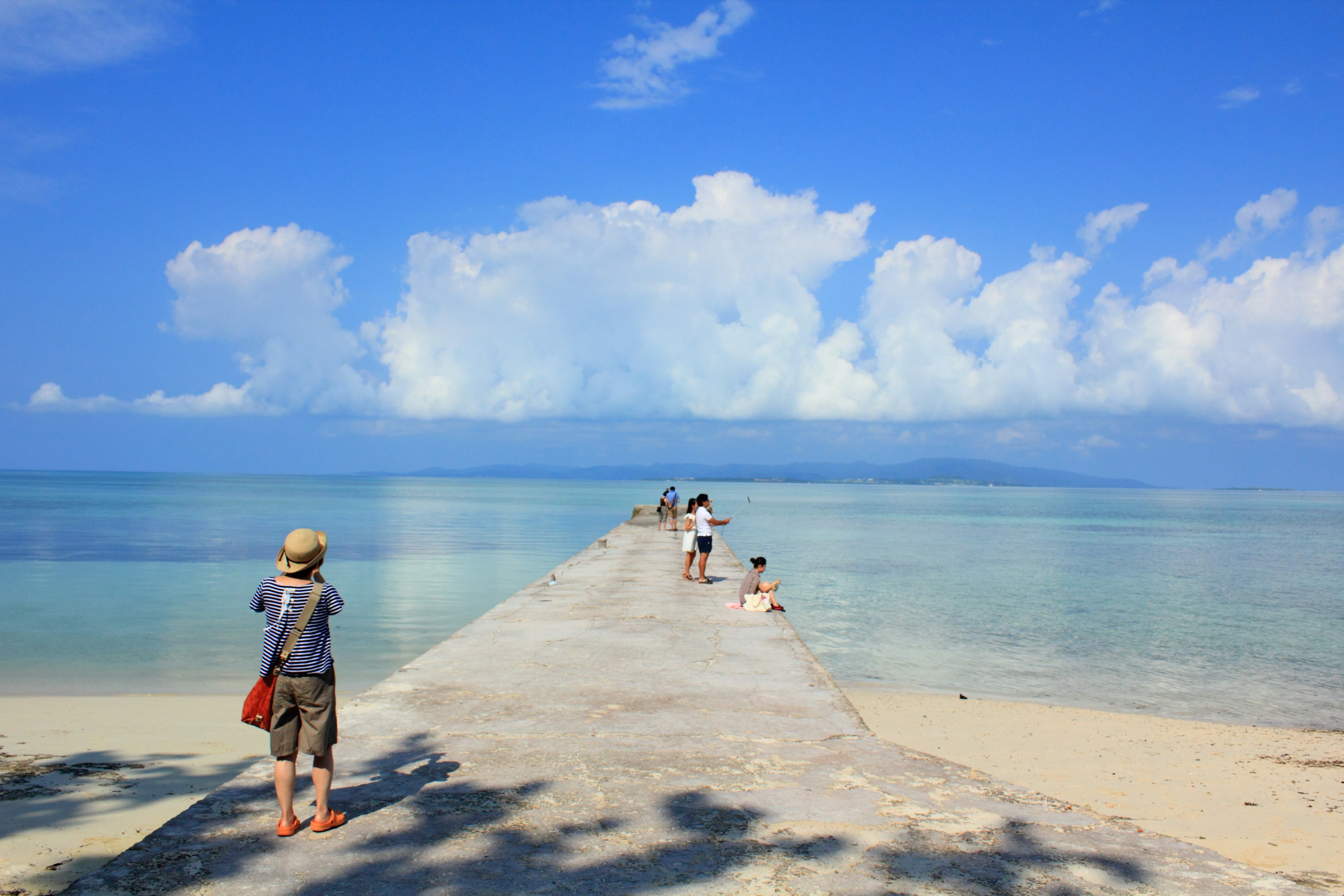 Personas caminando por un muelle que se extiende en un mar azul claro bajo un cielo brillante