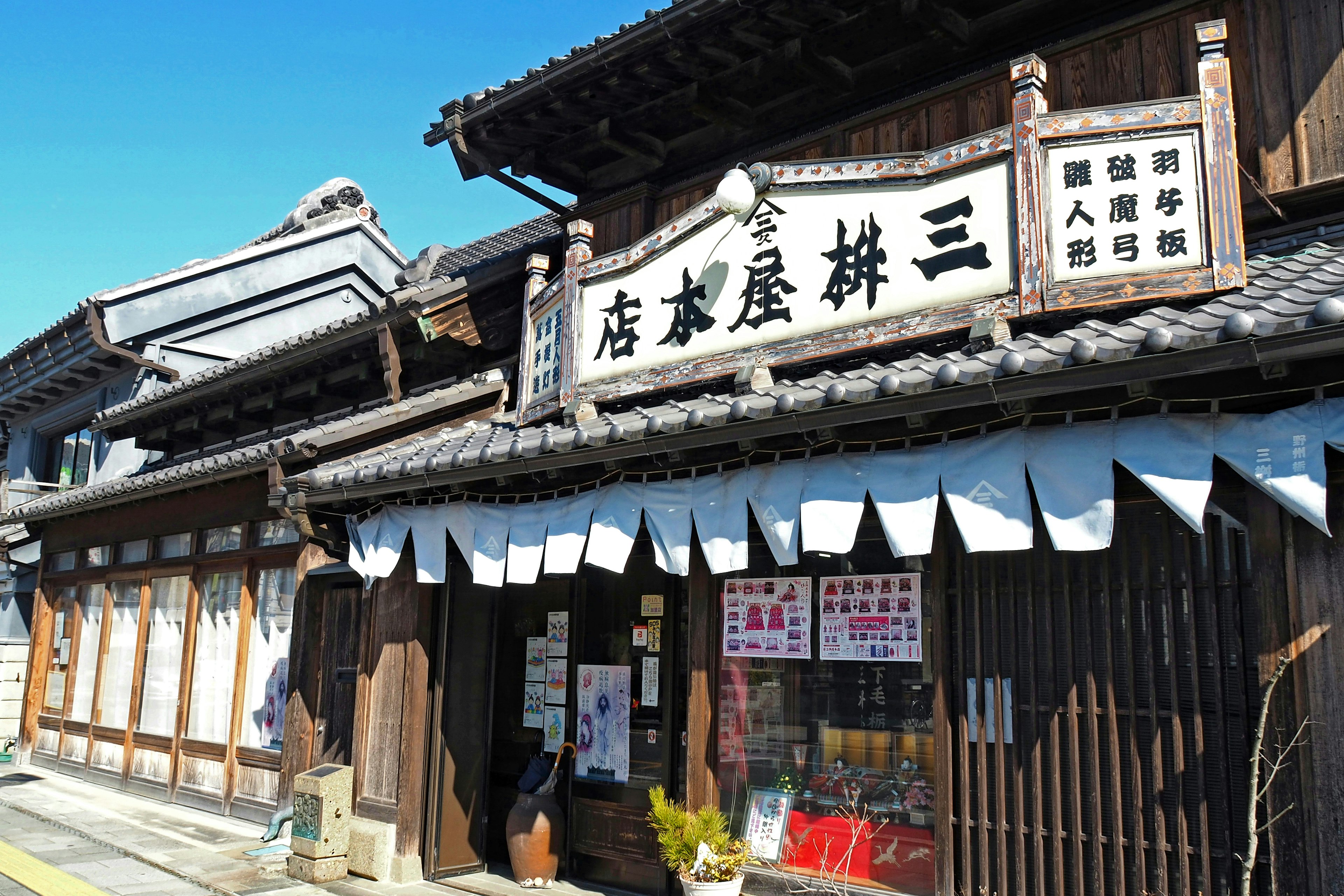 Eksterior toko tradisional Jepang dengan struktur kayu di bawah langit biru dengan papan kanji