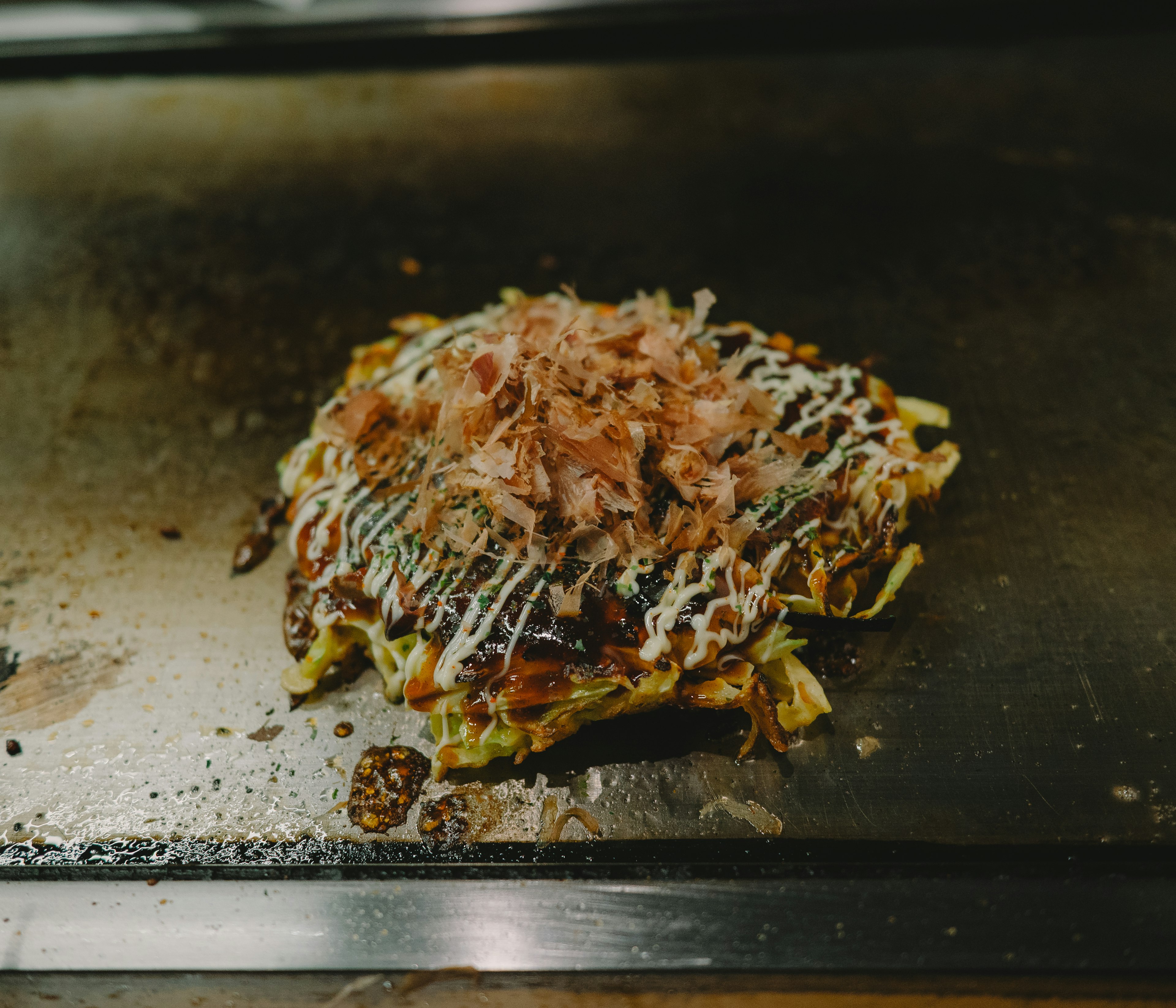 Okonomiyaki topped with bonito flakes and sauce on a griddle