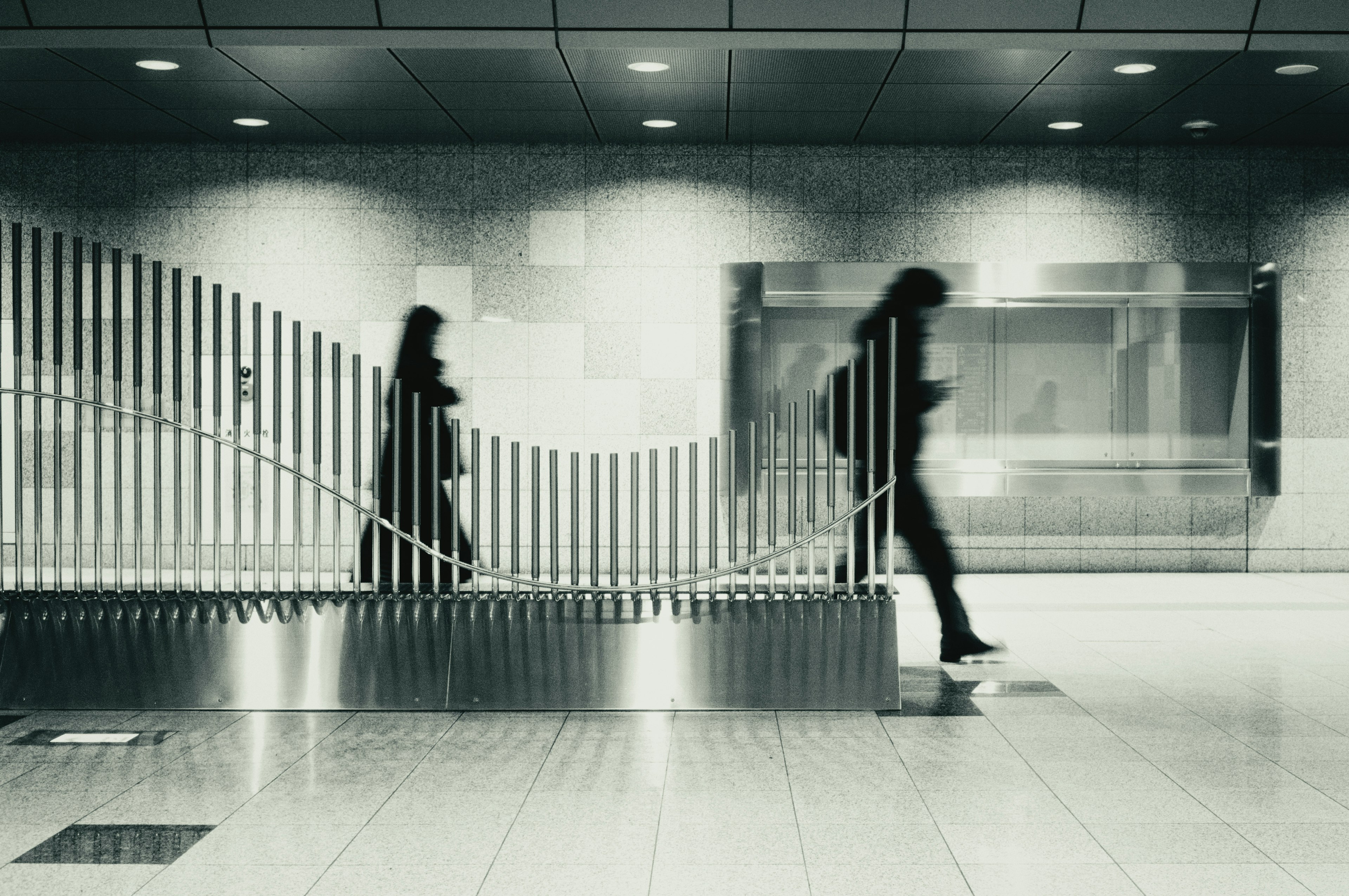 Silhouettes de deux personnes marchant dans un intérieur de gare monochrome avec un banc au design moderne