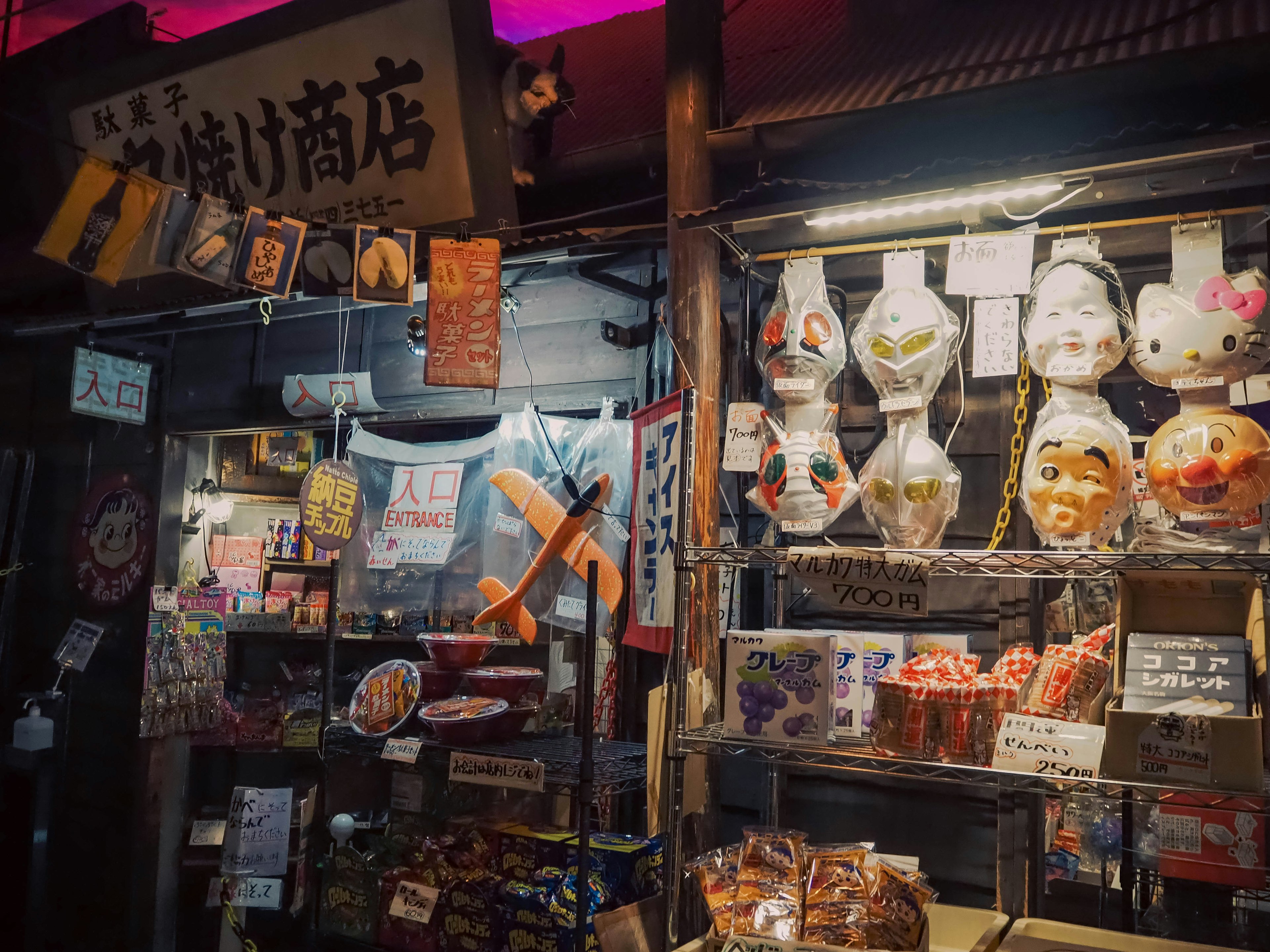 日本の伝統的なお菓子屋の外観が映る夜の風景で、色とりどりの飴細工やお面が並んでいる