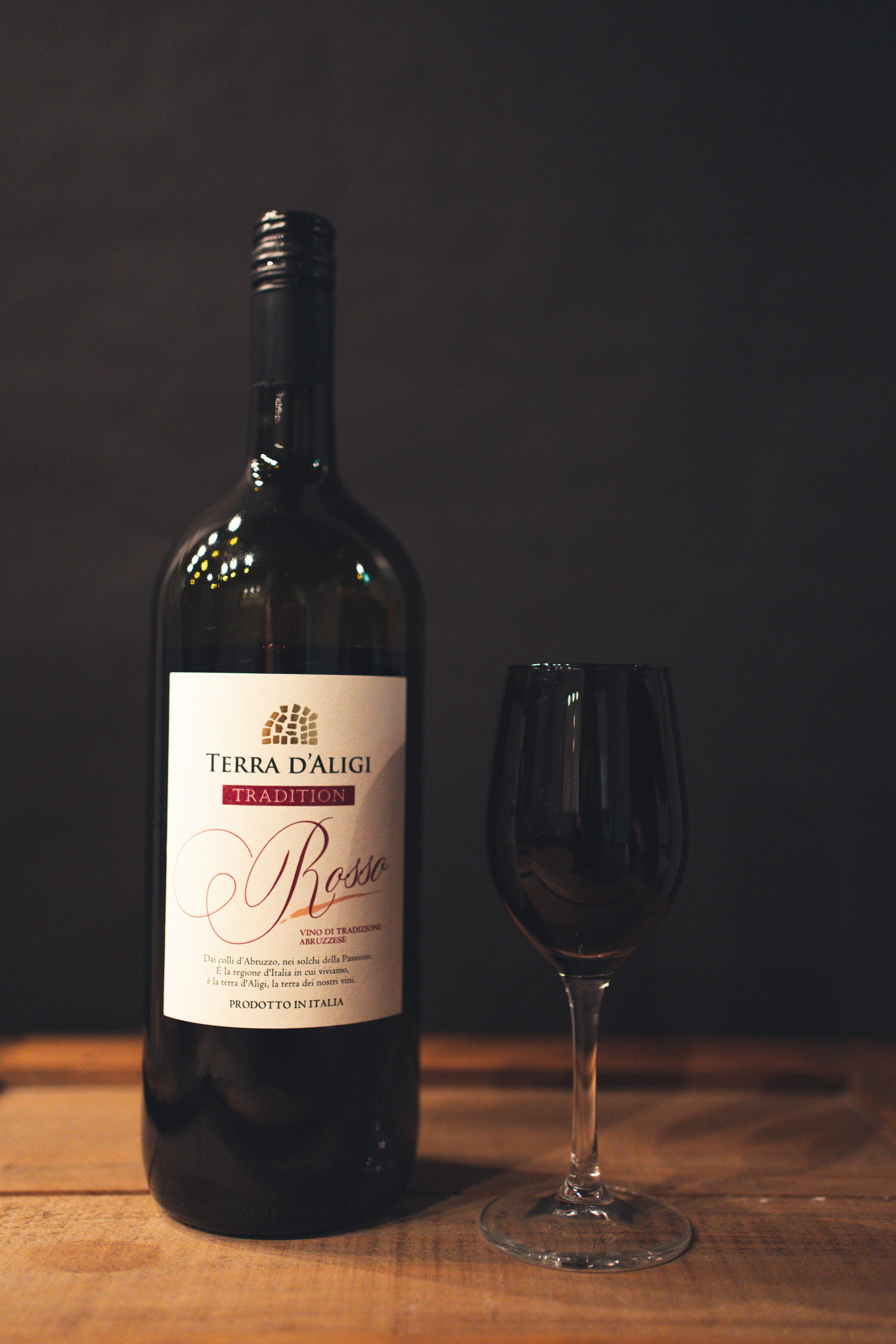 Large bottle of red wine next to a clear wine glass on a wooden table