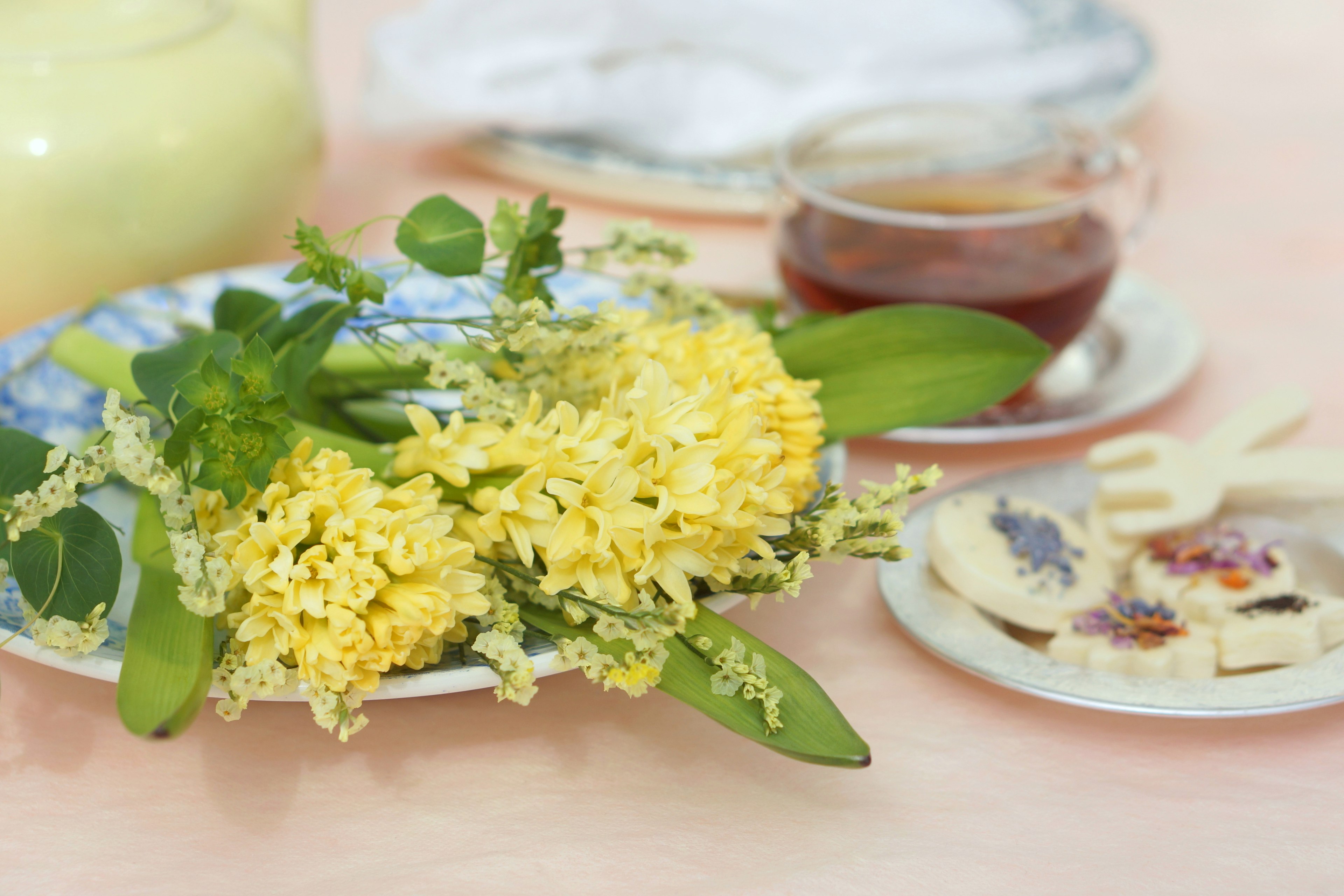 Eine ruhige Tischszene mit einem Teller gelber Blumen und grünen Blättern neben einer Tasse Tee und Keksen