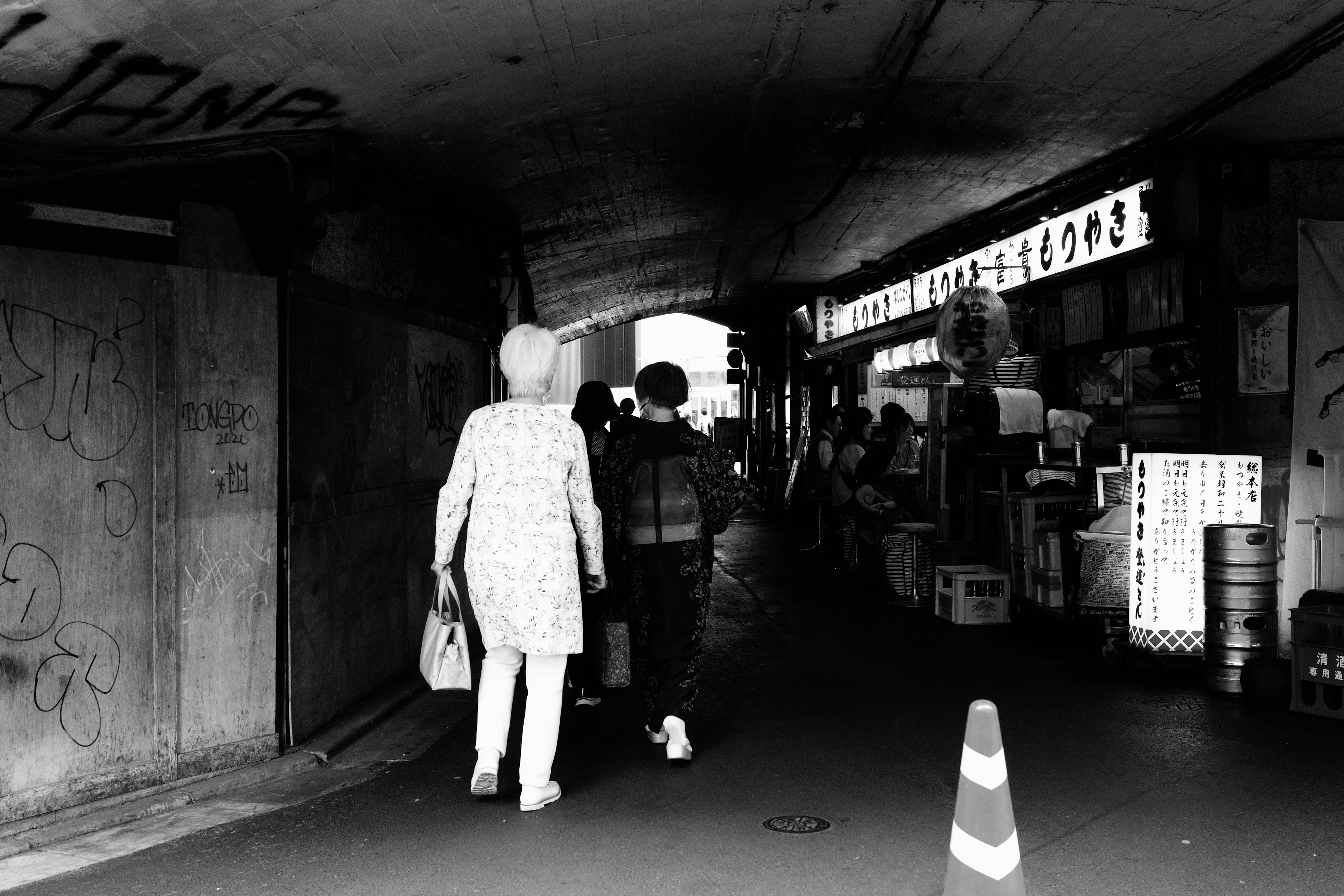 Siluetas de dos personas caminando bajo un túnel en blanco y negro con tiendas alrededor