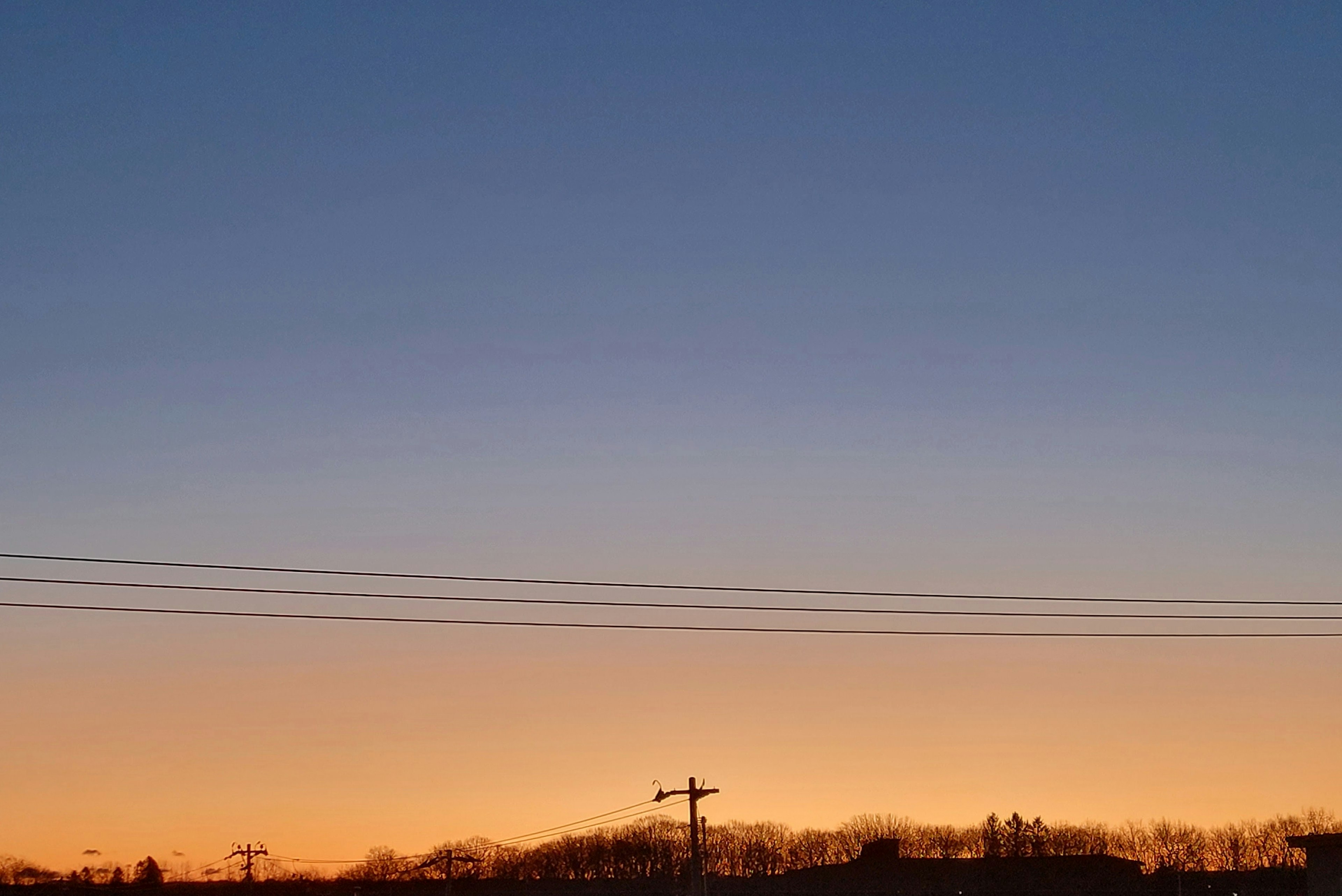 Ein Farbverlauf des Himmels von Blau zu Orange bei Sonnenuntergang mit Stromleitungen und silhouettierten Hügeln