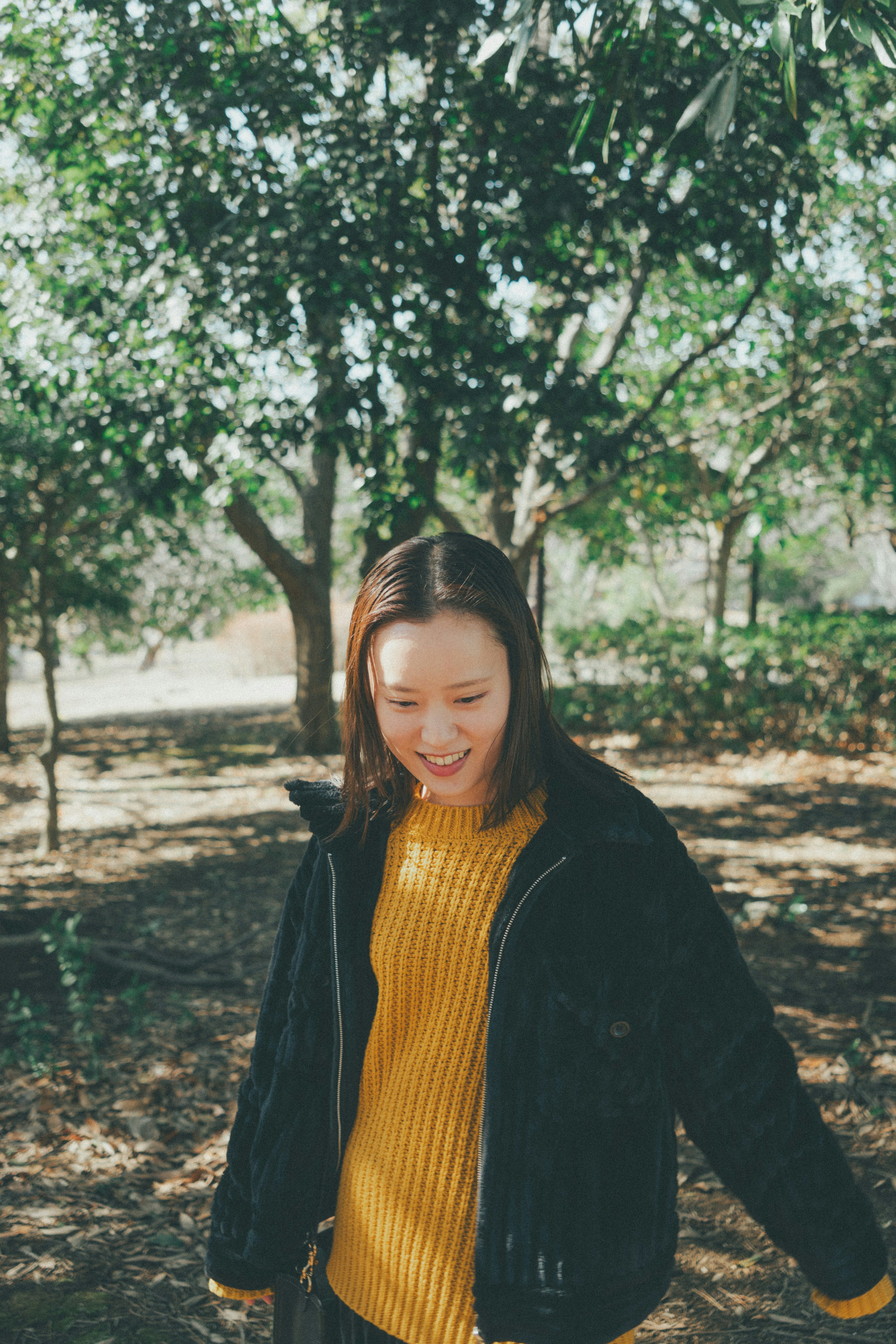 Une femme souriante en pull jaune et veste noire se tenant parmi des arbres verts