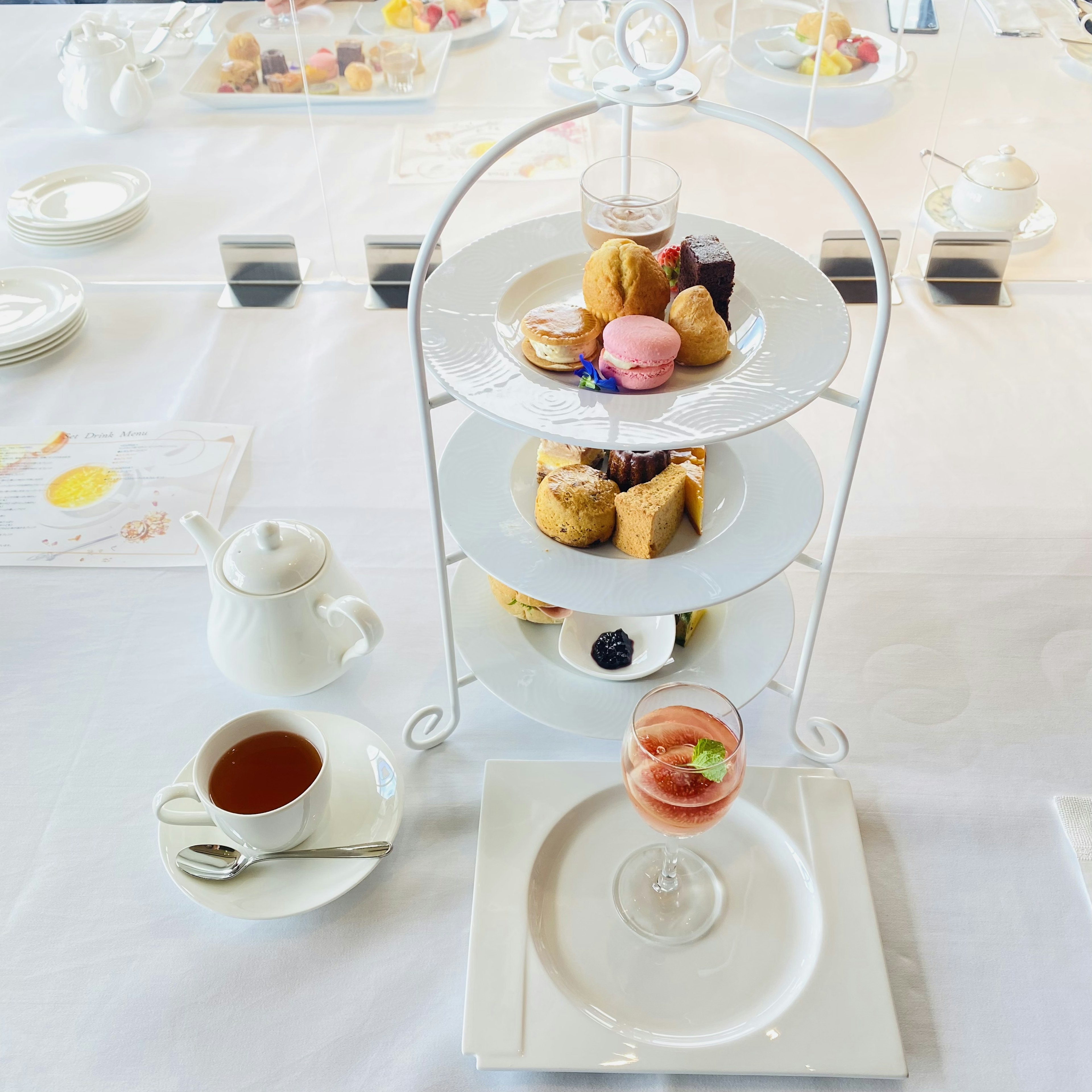 Un portatè a tre piani pieno di dolci colorati su una tovaglia bianca accompagnato da una tazza di tè e un piccolo bicchiere di dessert