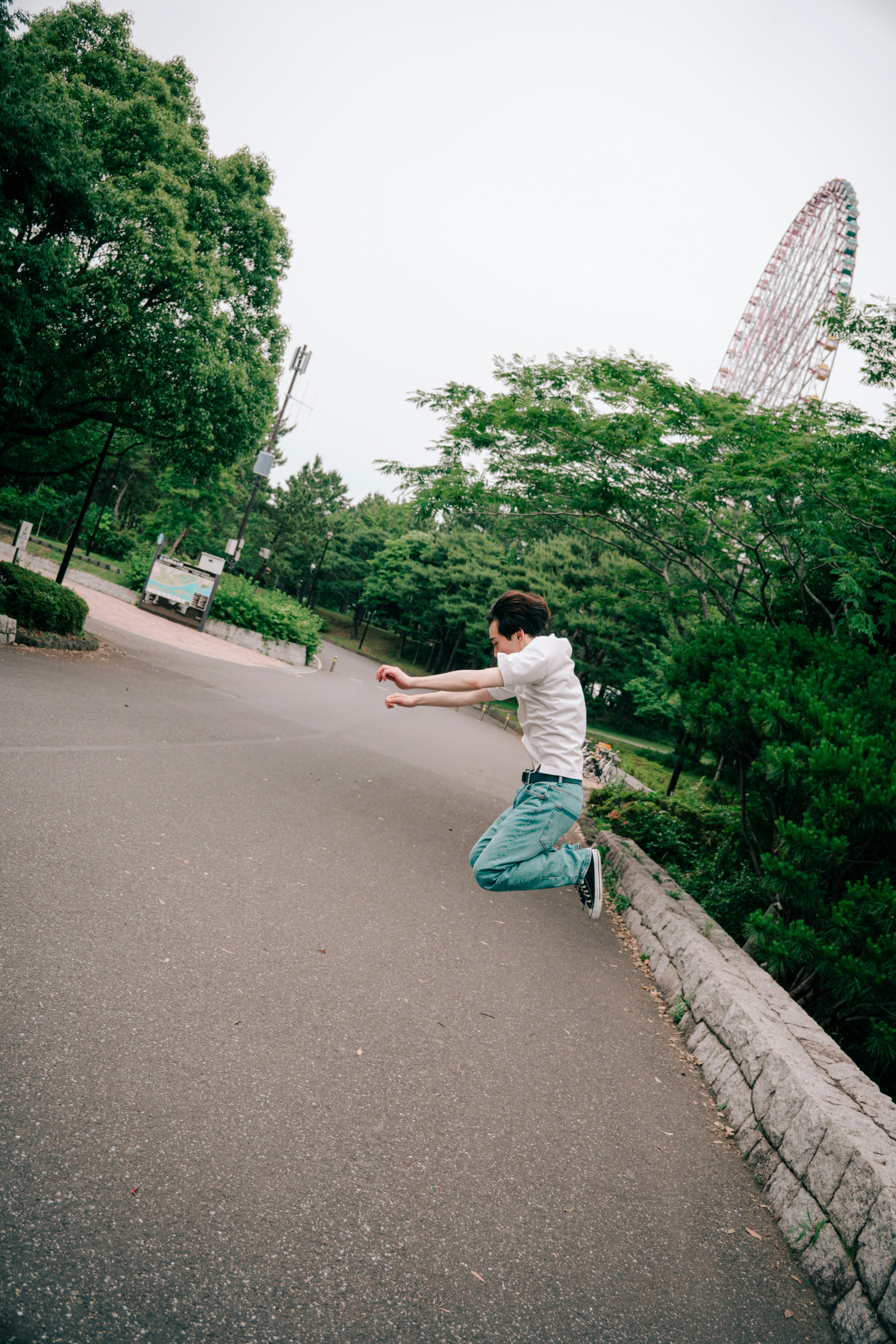 Young person jumping on a park path surrounded by green trees and a Ferris wheel in the background