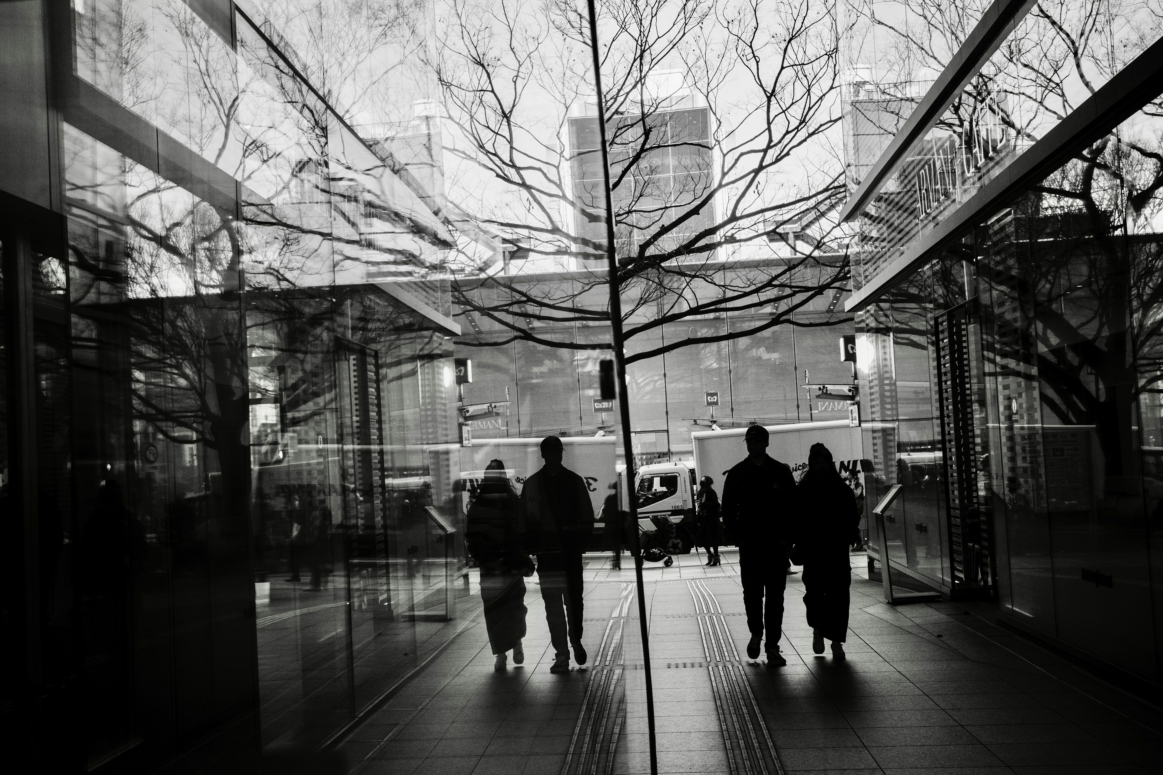 View from inside a glass building showing people walking outside silhouettes of trees reflected