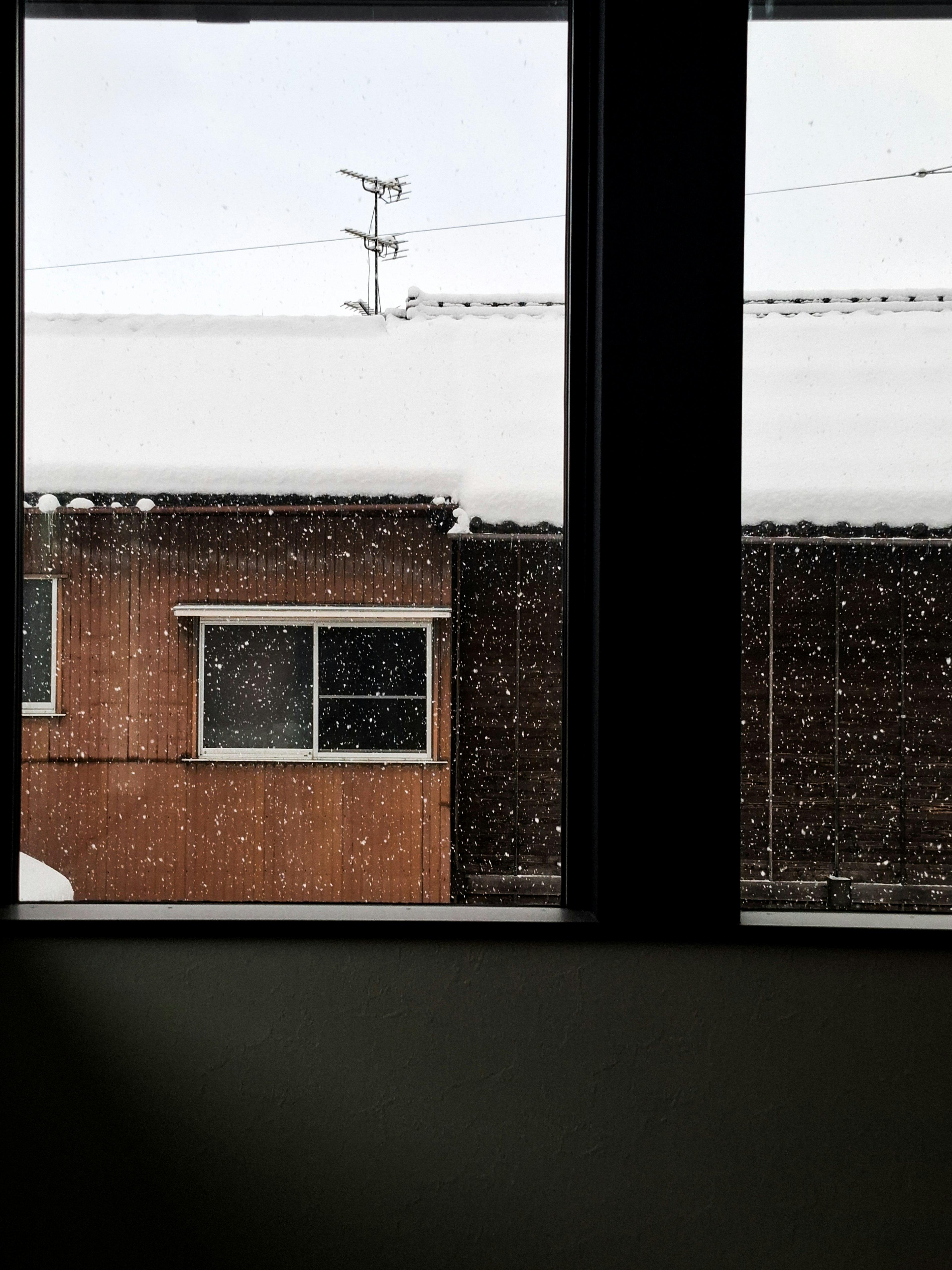 窗外飄落的雪，視野中有棕色房屋和積雪覆蓋的屋頂