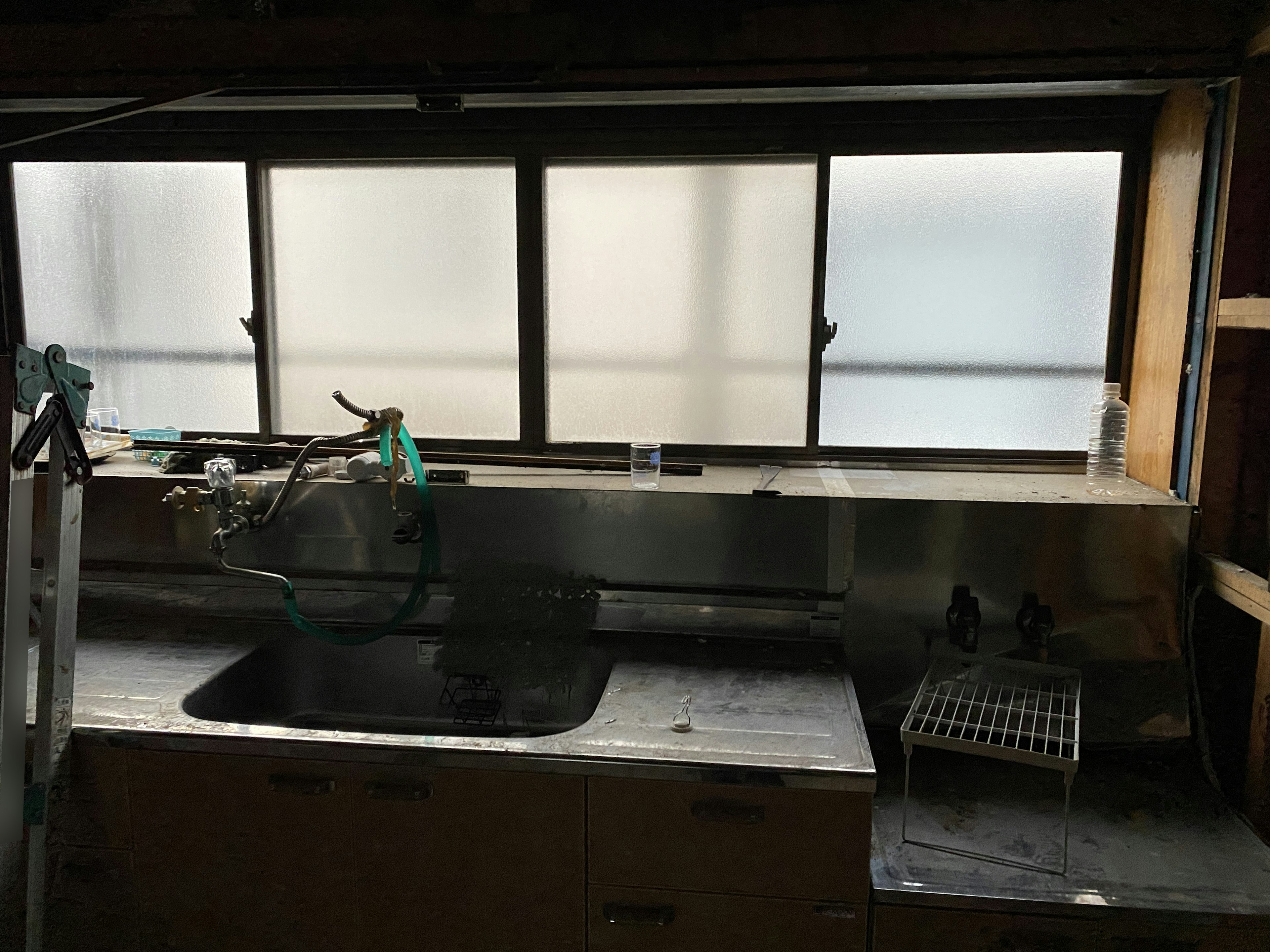 Interior scene of a simple kitchen with windows featuring a stainless steel sink and cooking utensils