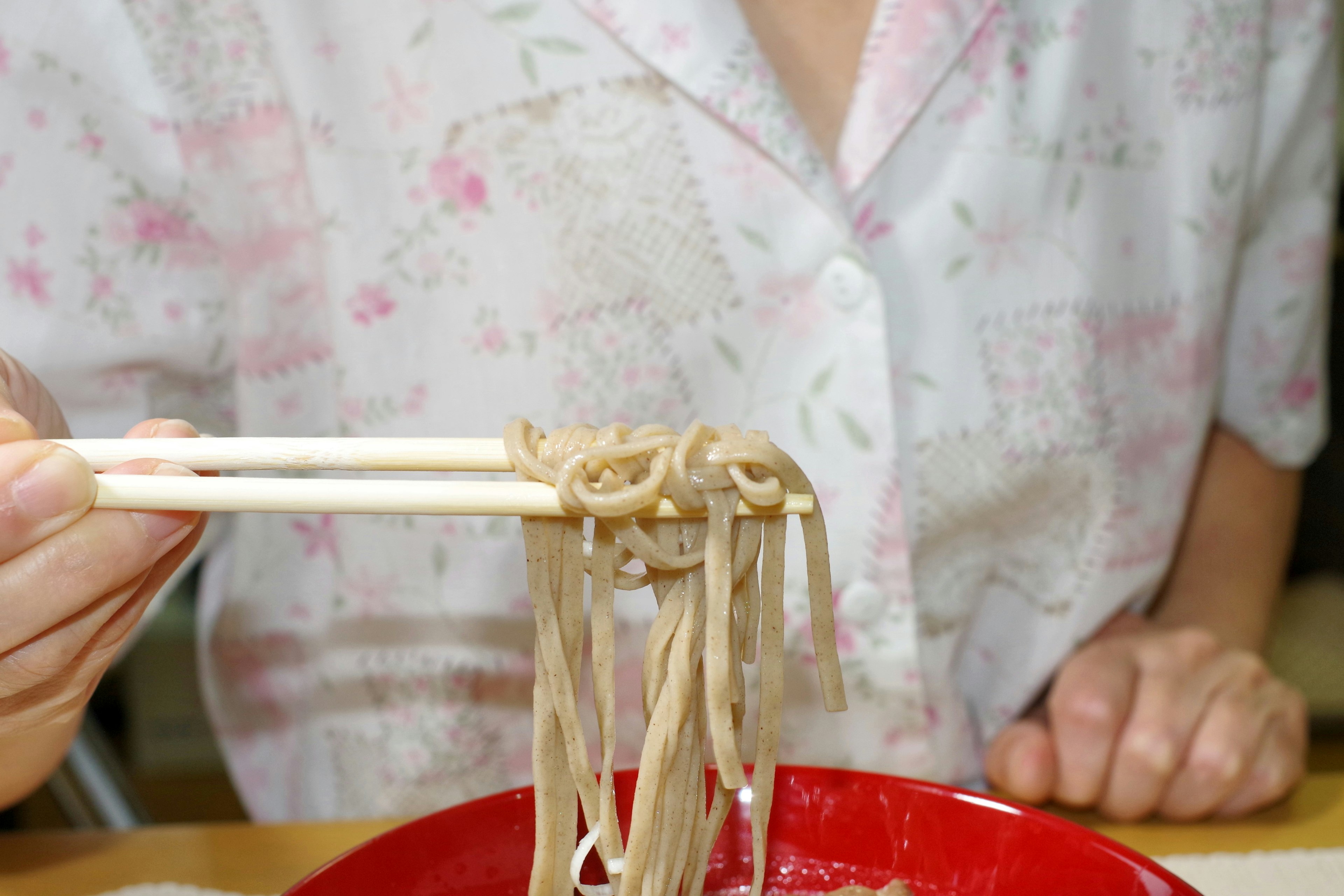 A woman holding soba noodles with chopsticks in a red bowl