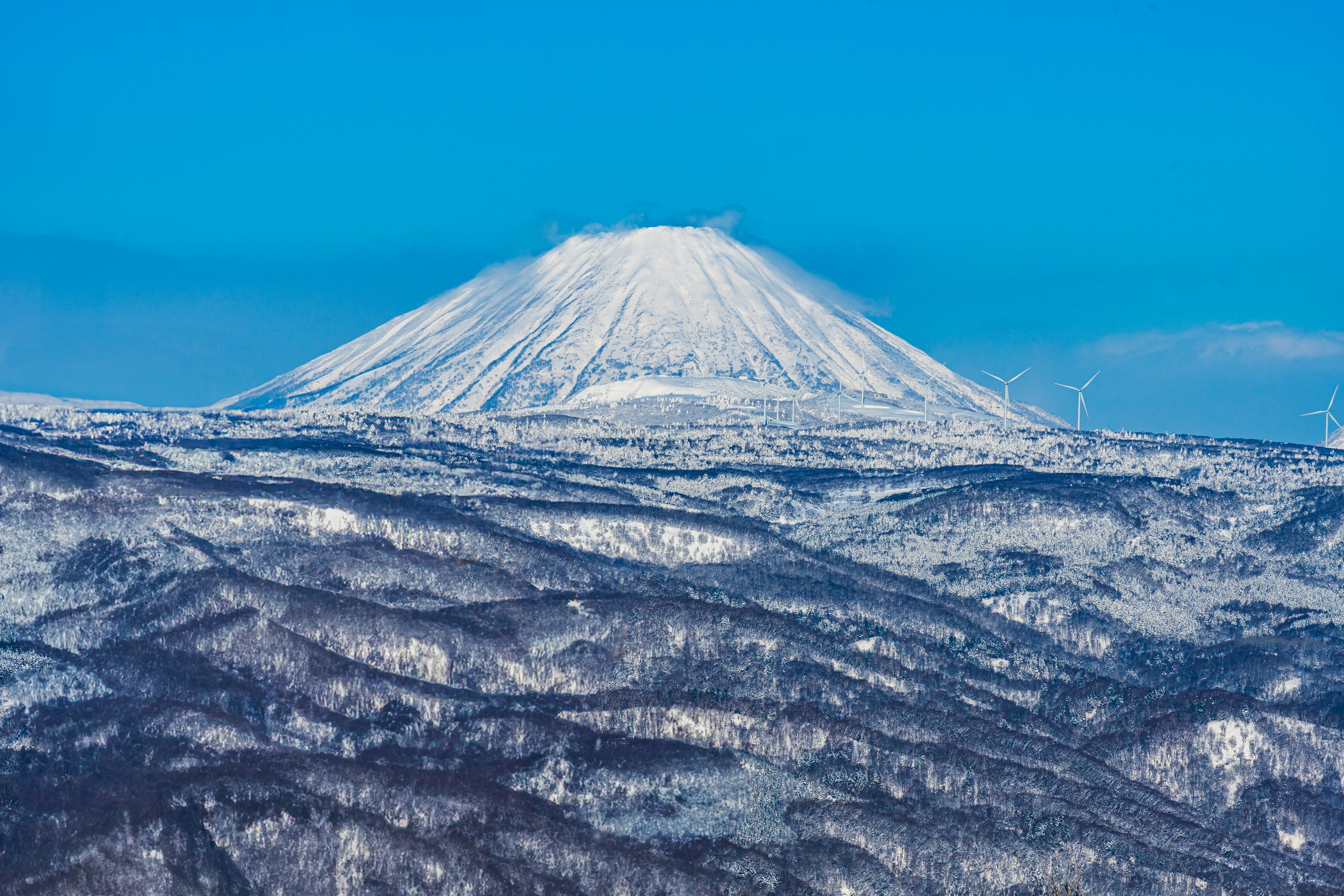 Paisaje montañoso cubierto de nieve bajo un cielo azul claro con turbinas eólicas visibles