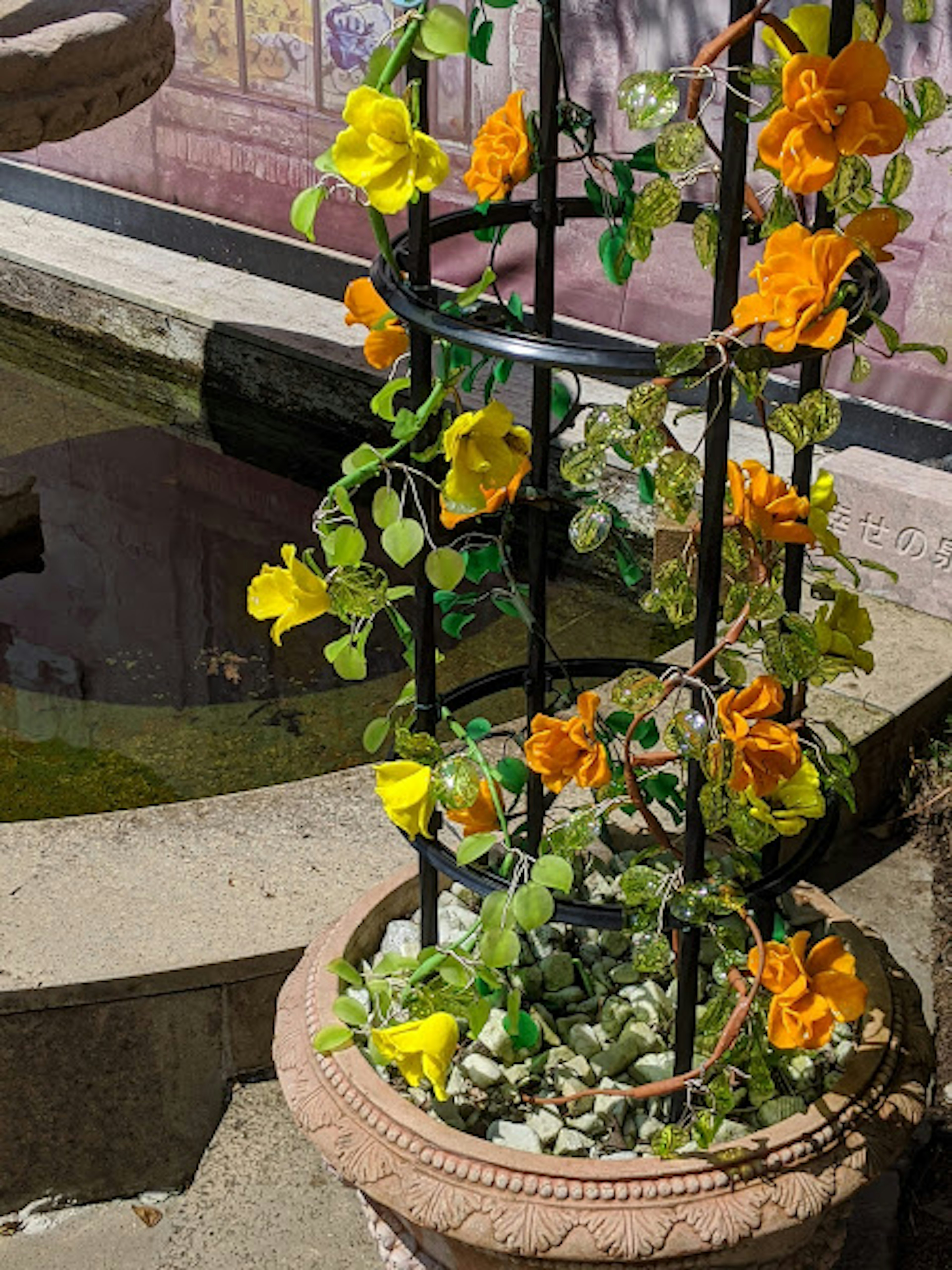 Planta en maceta con flores naranjas y amarillas trepando por un enrejado negro cerca de un estanque