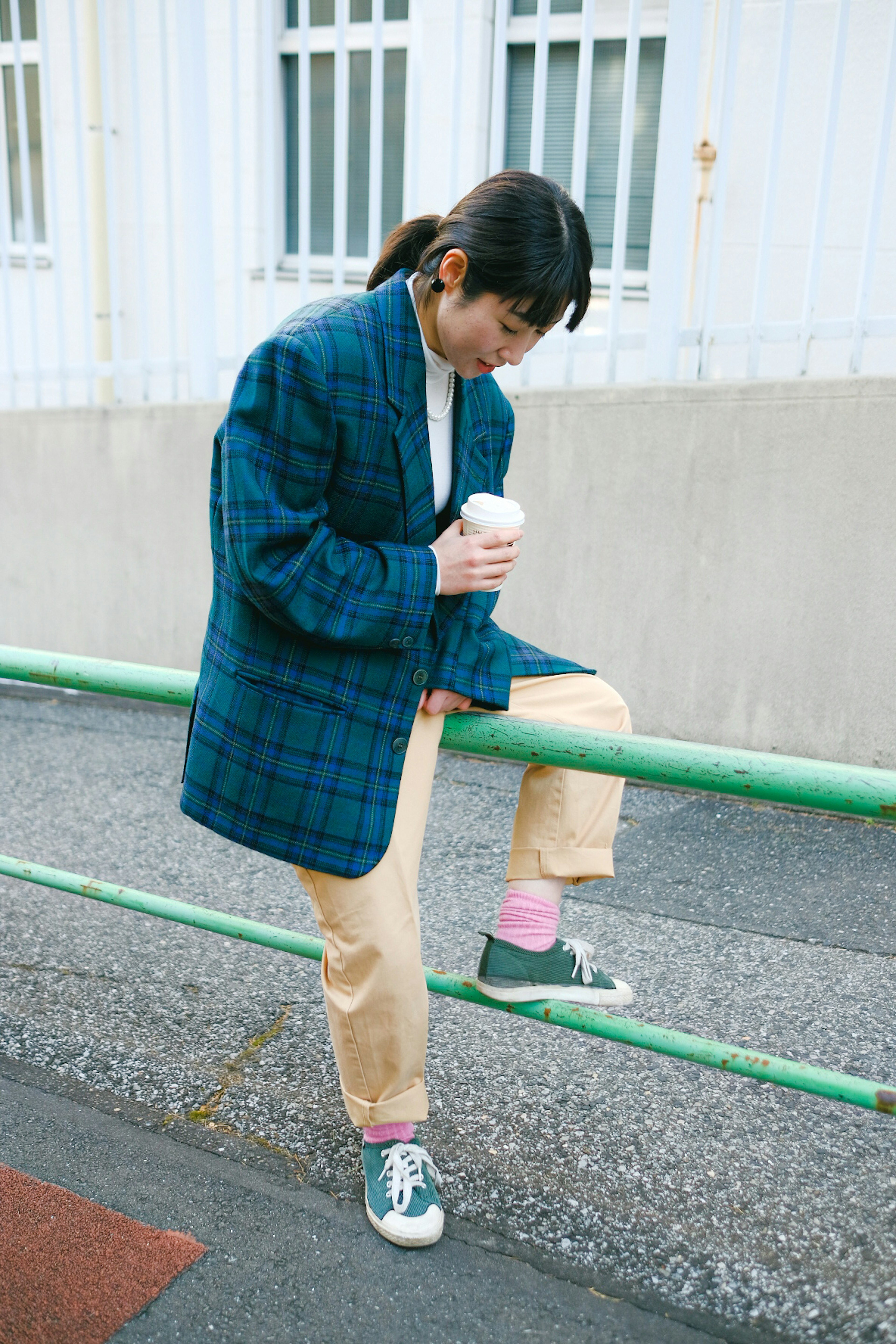 A woman wearing a checkered jacket leaning against a green railing holding a coffee cup