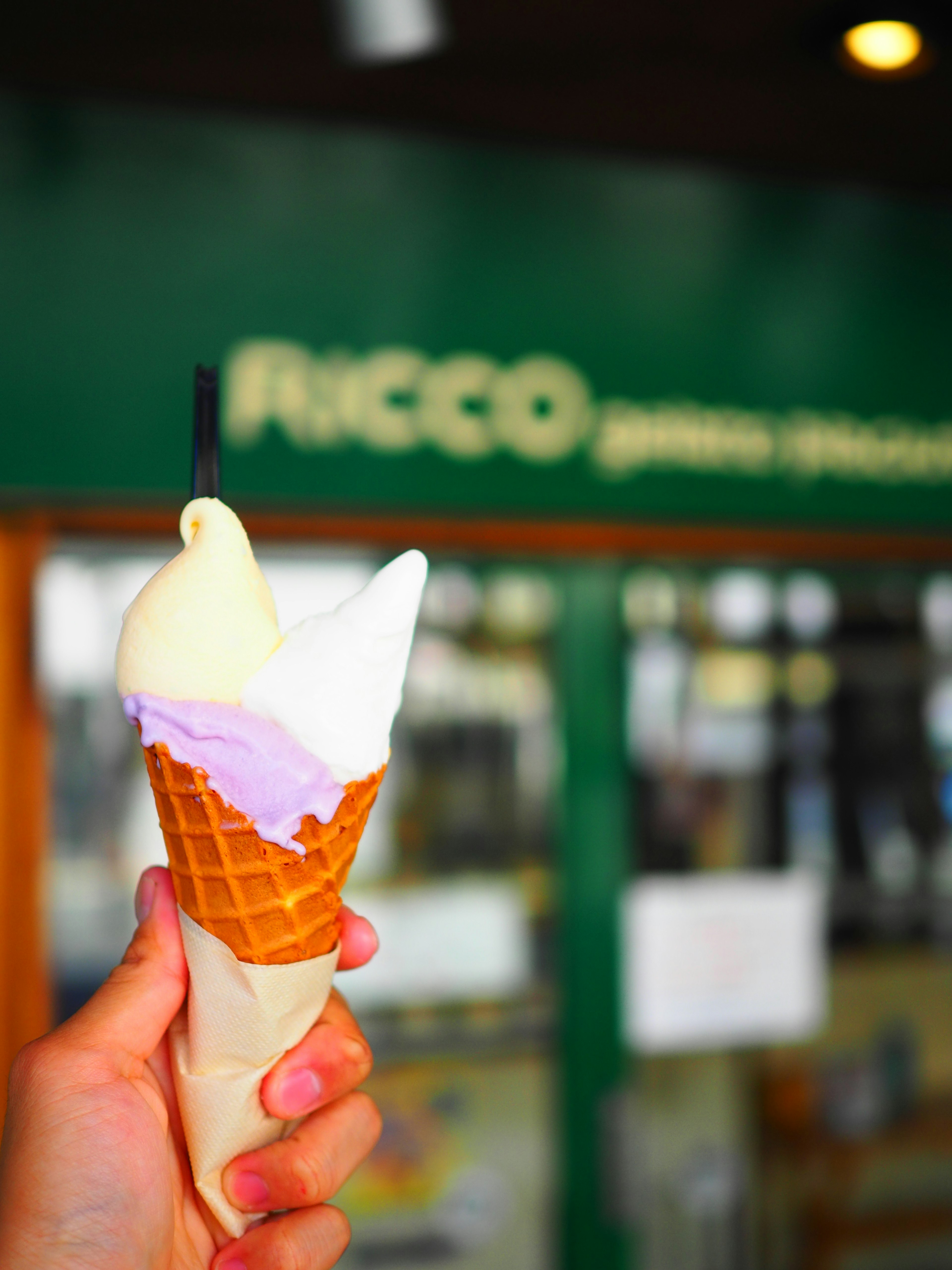 Cono de helado de vainilla y sabor púrpura sostenido a mano nombre de la tienda RICCO al fondo