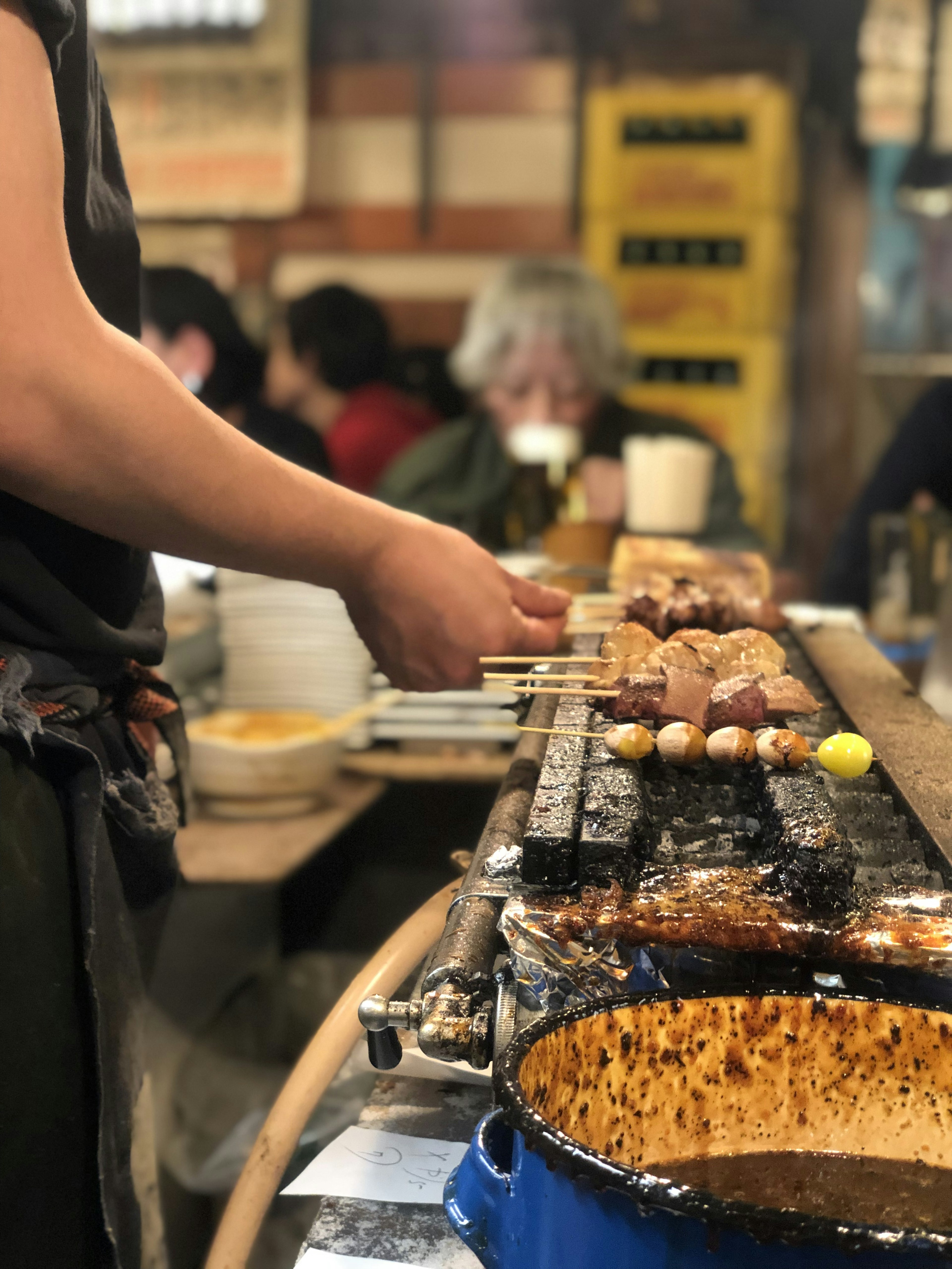 Cook grilling yakitori with skewers in a bustling eatery filled with patrons