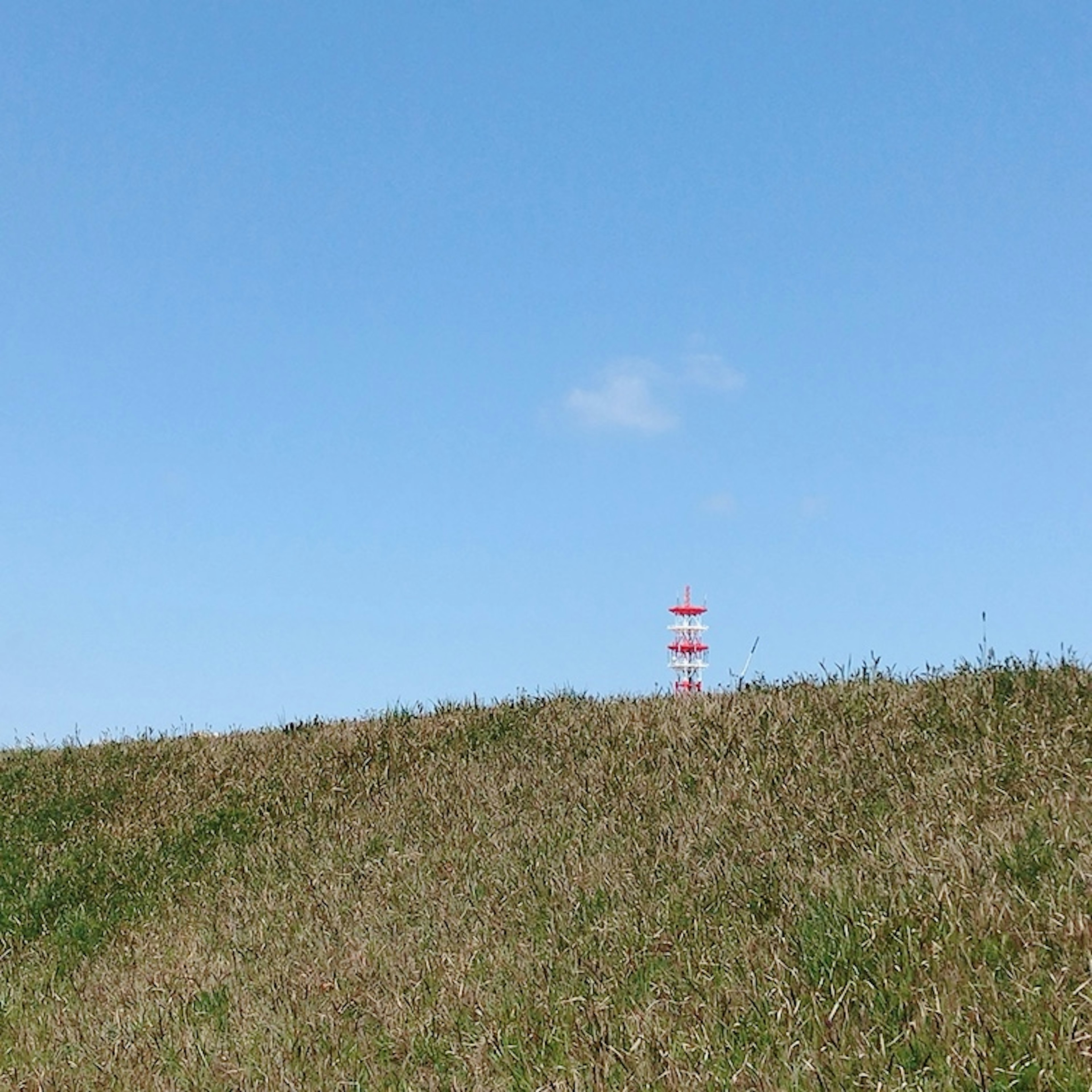 青空と緑の丘に赤白の塔が立っている風景