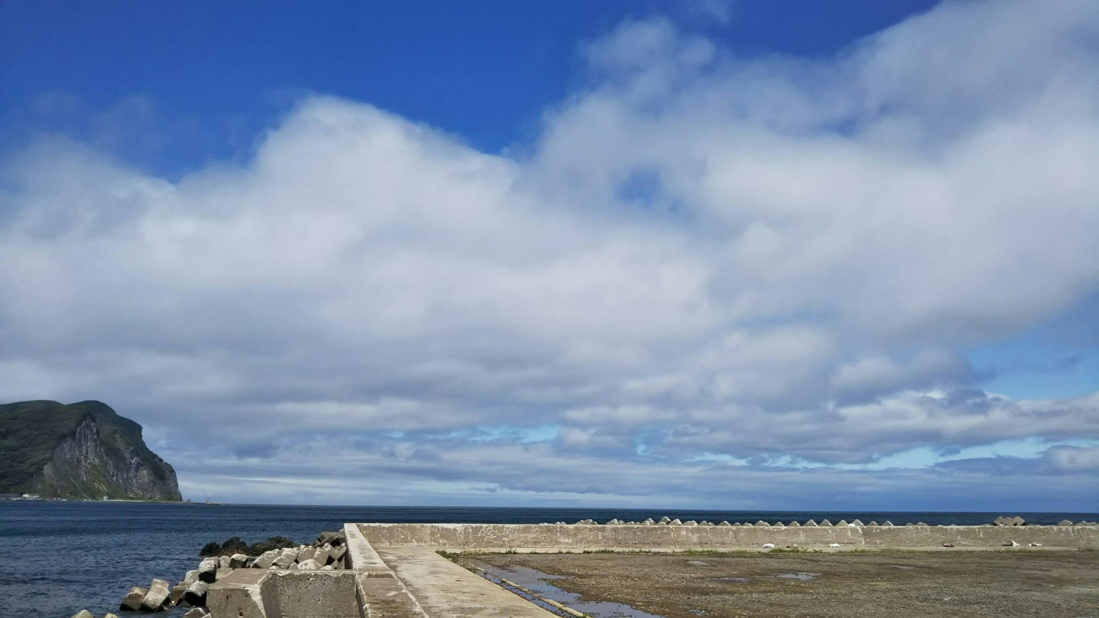 Küstenansicht mit blauem Himmel und weißen Wolken Pier-ähnliche Struktur, die ins Meer ragt