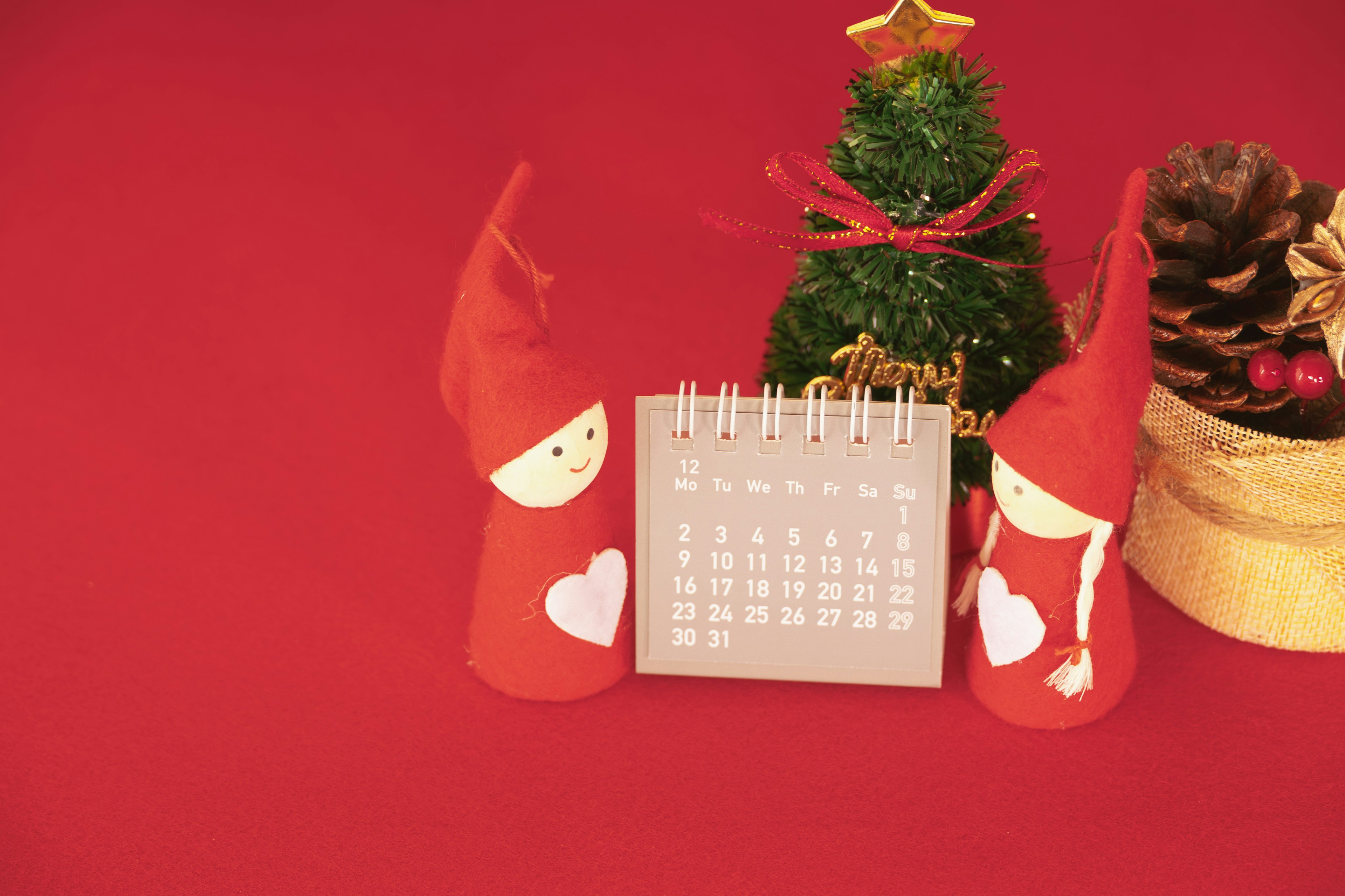 Immagine di due bambole con cappello rosso e un piccolo albero di Natale con un calendario su uno sfondo rosso