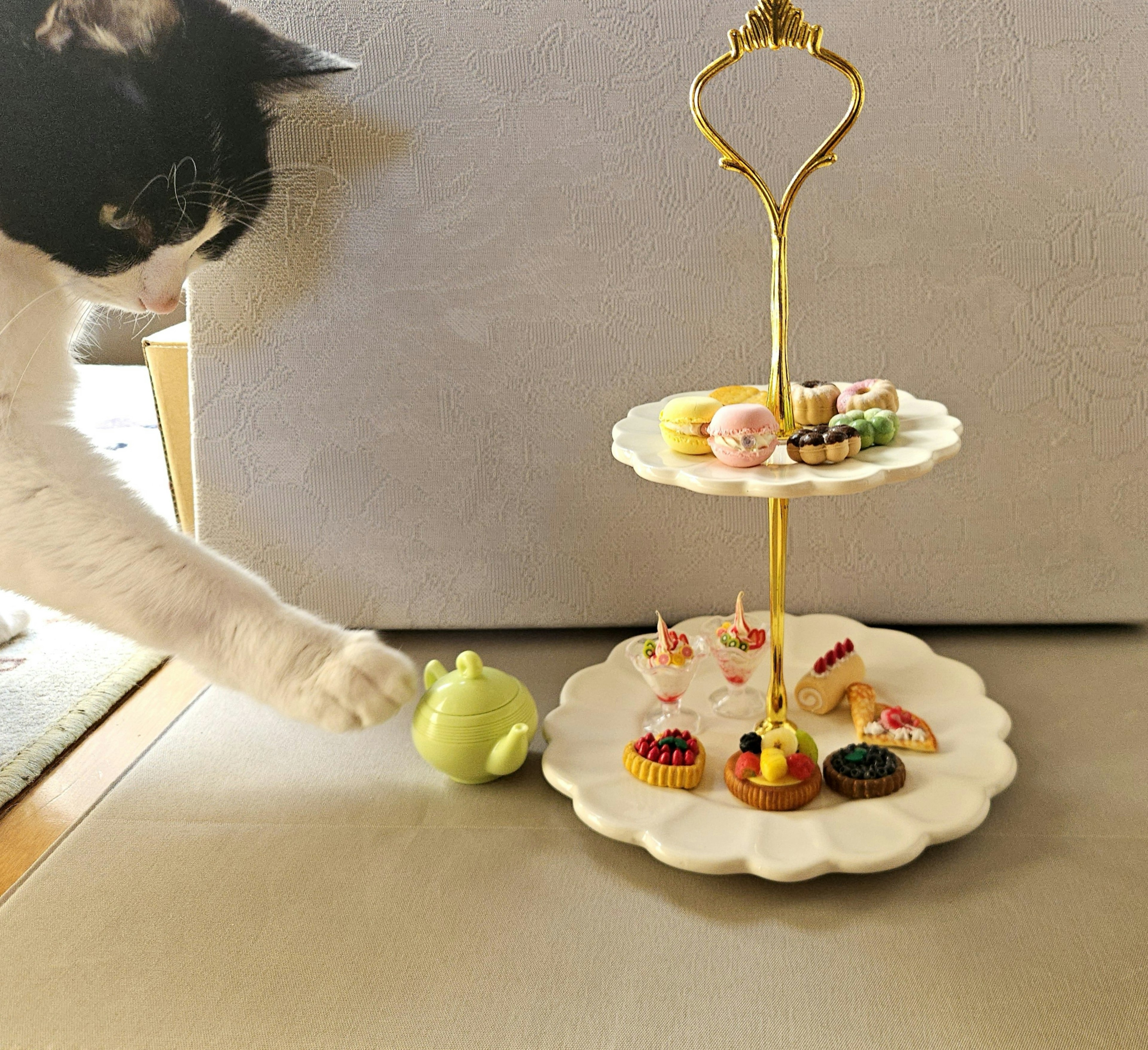 A cat reaching for a small green object near a tiered cake stand with colorful desserts