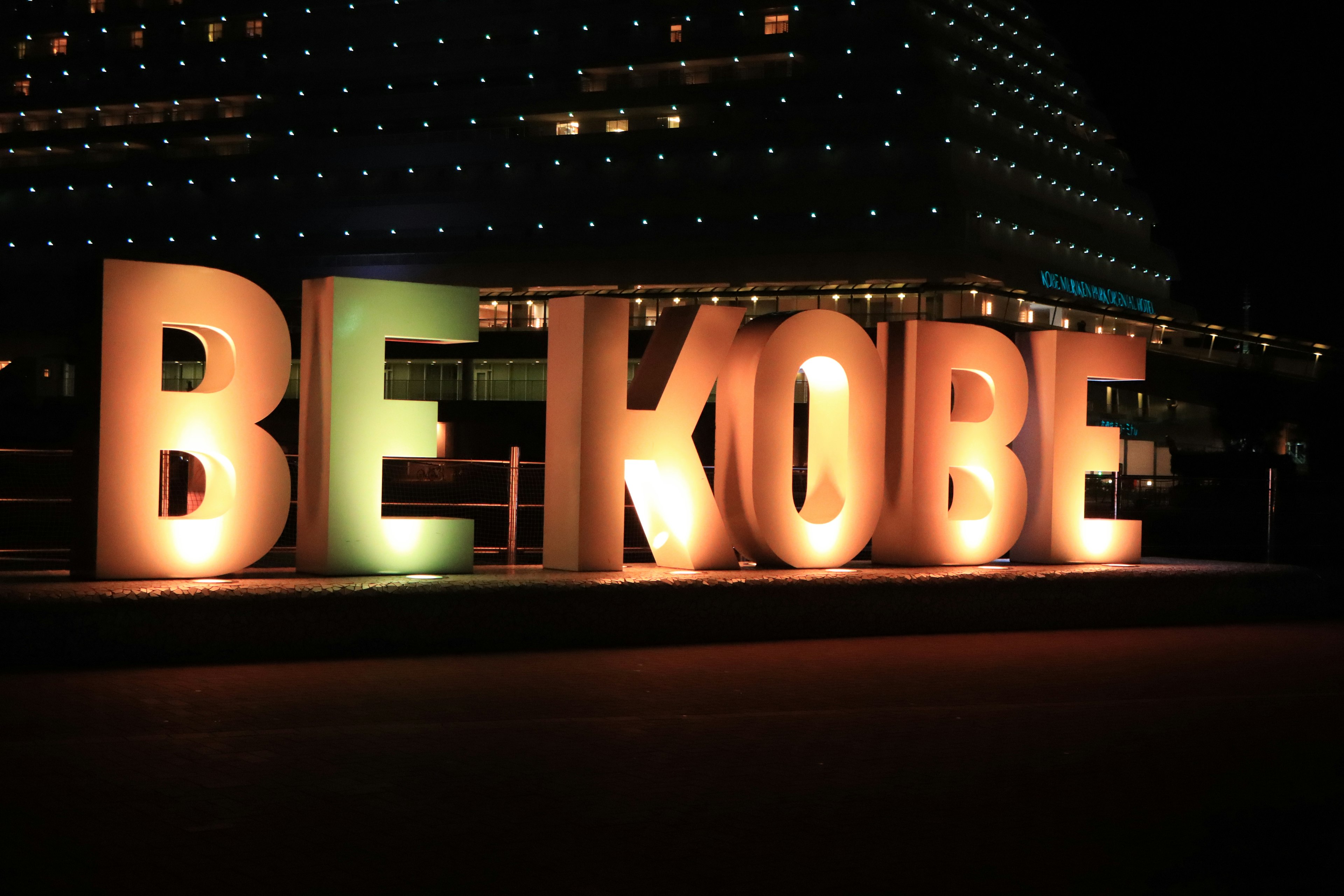 Large illuminated letters spelling BE KOBE against a nighttime backdrop