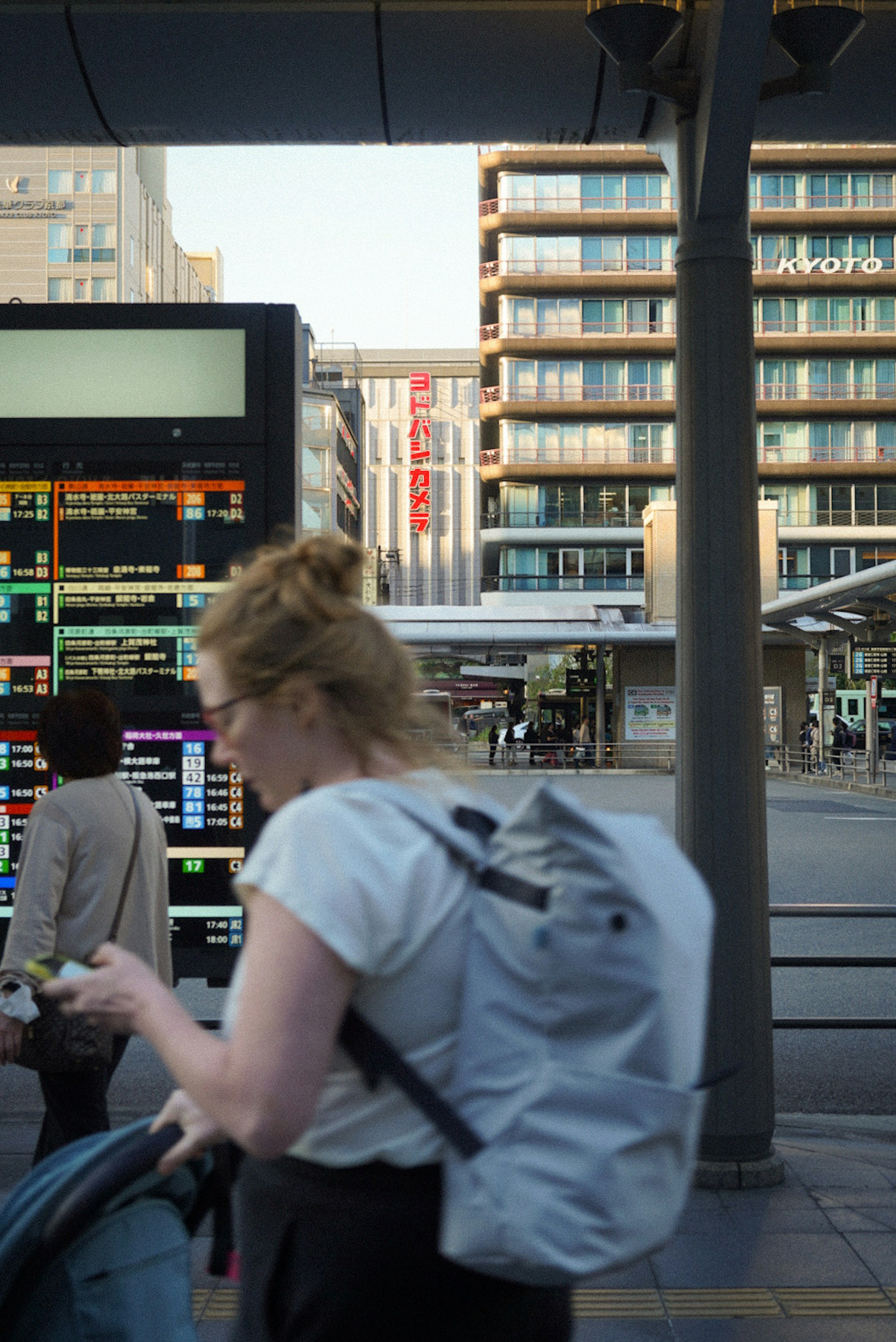 Urban scene featuring a station information board and buildings a woman using a smartphone