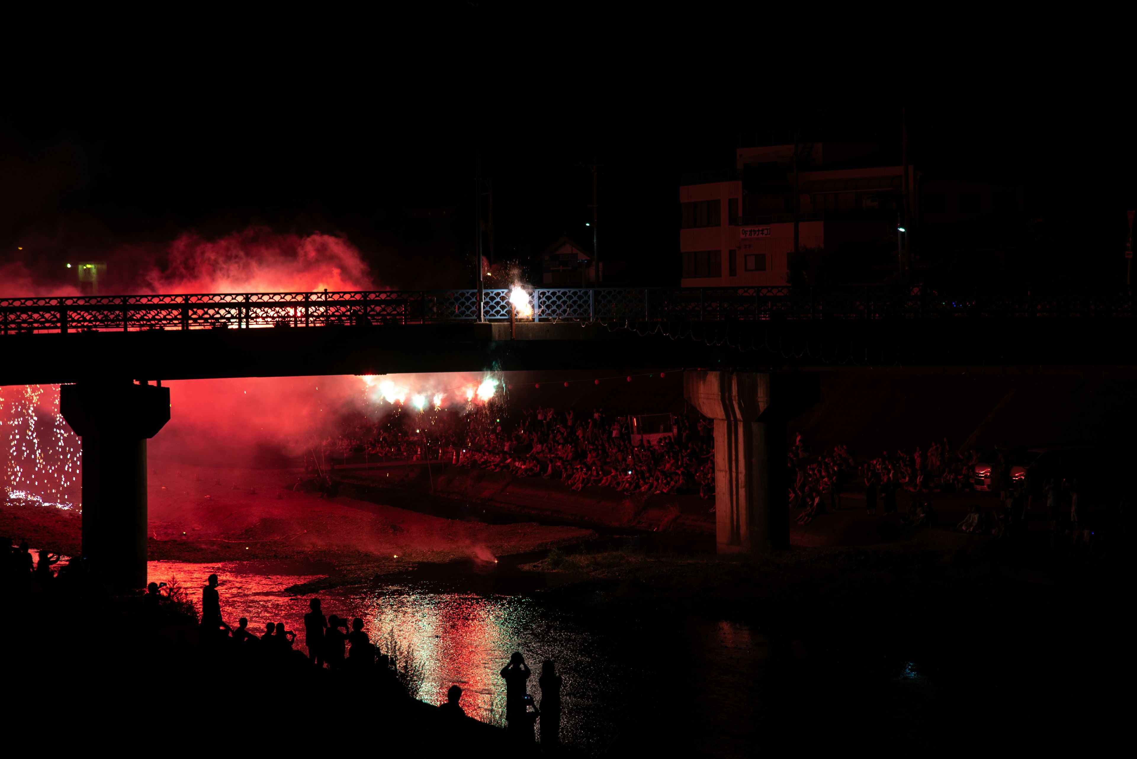 Espectáculo de fuegos artificiales nocturno con humo rojo y espectadores reunidos junto al río