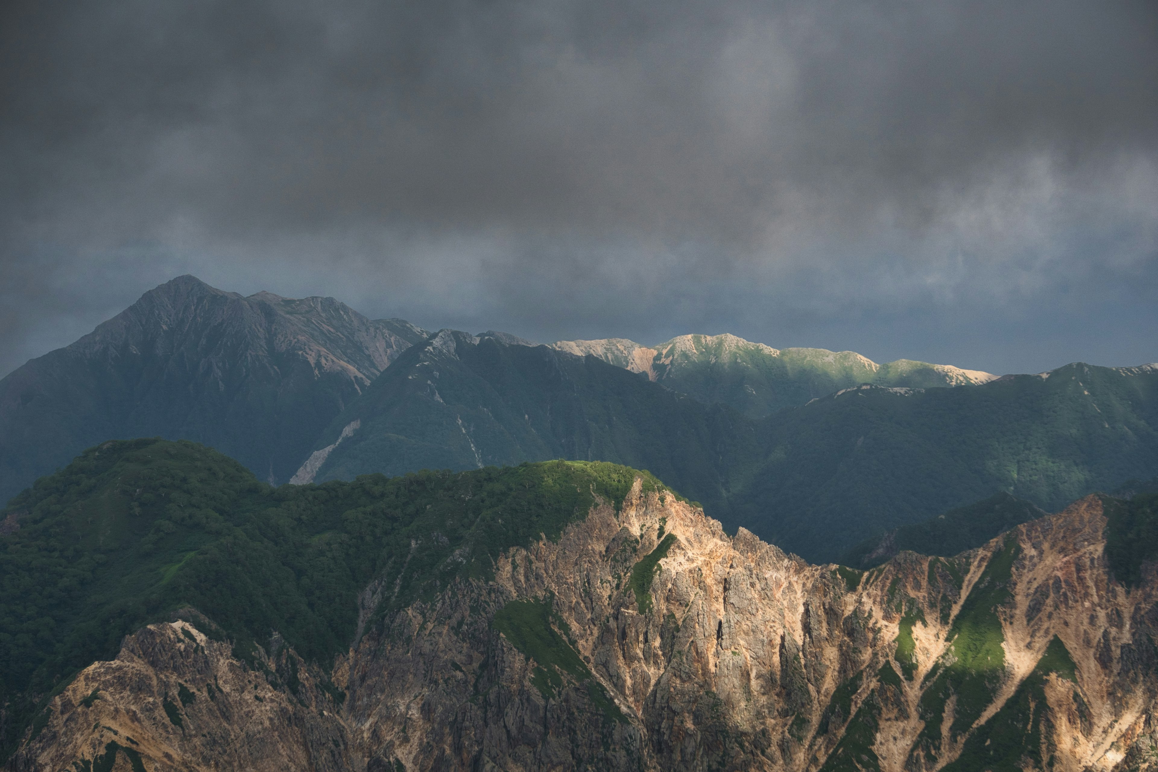 Paisaje montañoso bajo nubes oscuras con laderas verdes y picos rocosos brillantes