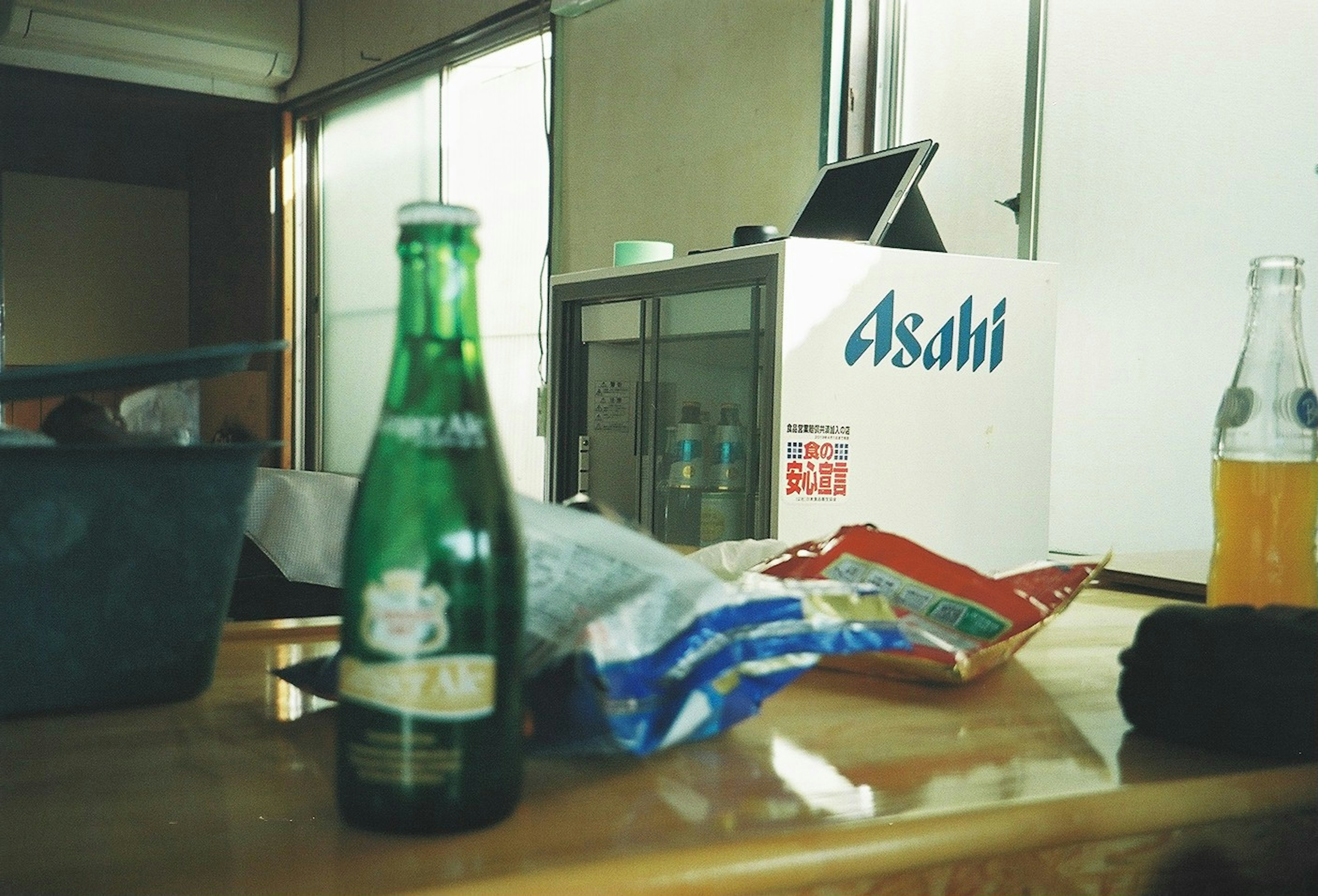 Una botella de cerveza verde y una bolsa de aperitivos sobre una mesa con un refrigerador Asahi al fondo