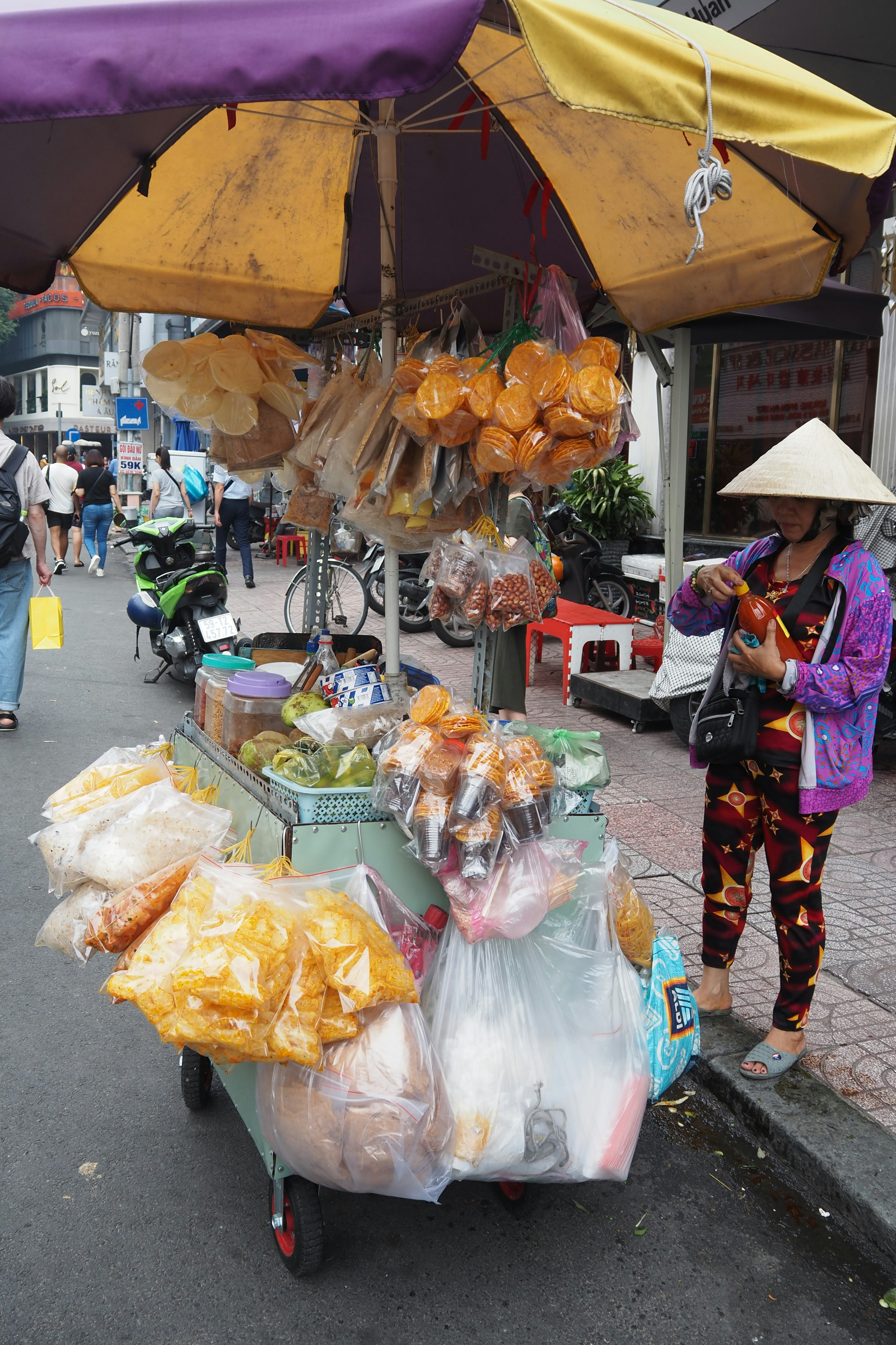 รถเข็นขายของข้างถนนสีสันสดใสที่แสดงผลไม้และขนมหลากหลายชนิดพร้อมกับผู้หญิงในชุดประจำชาติ