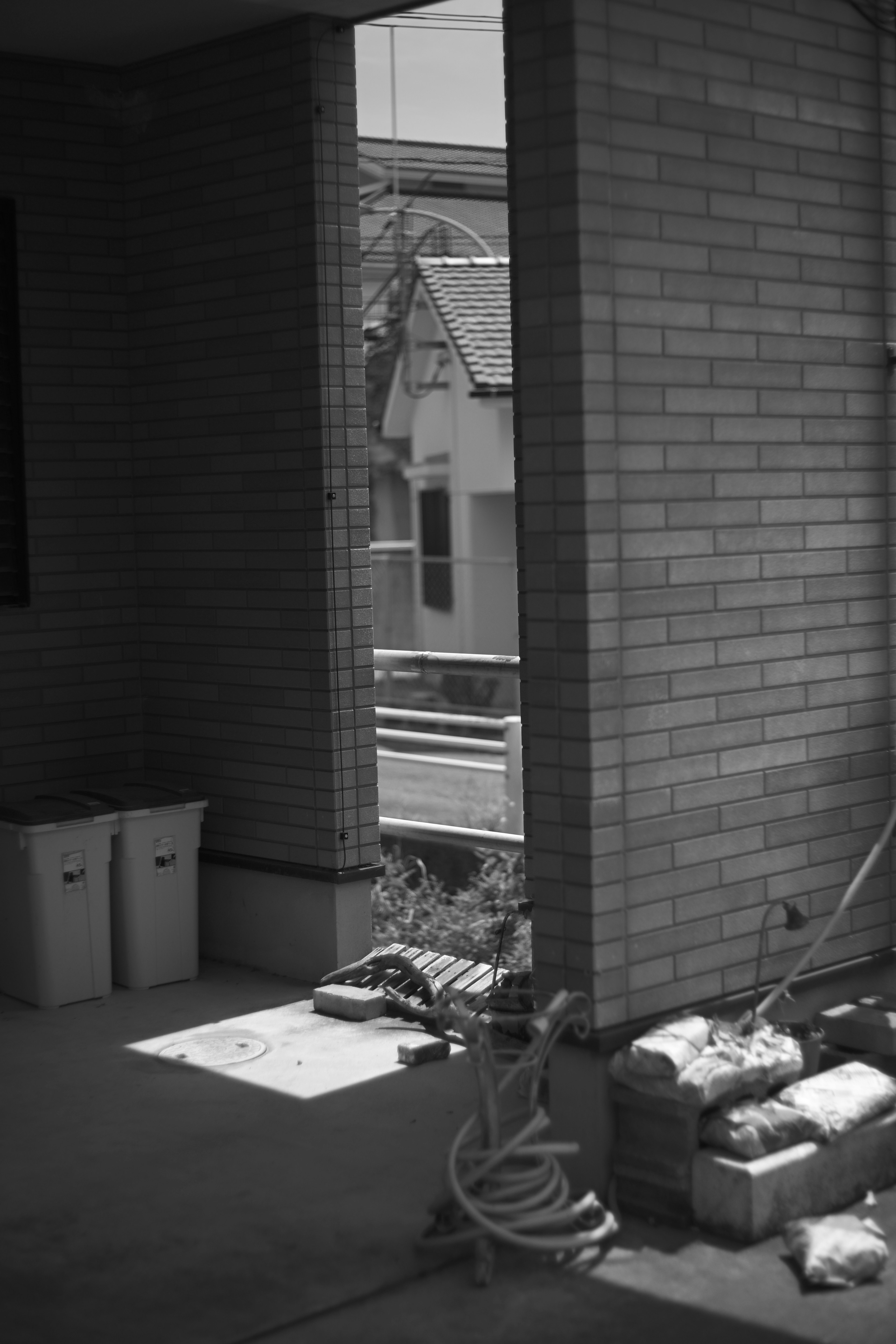 Black and white image showing the view from inside a building with trash bins and construction materials nearby