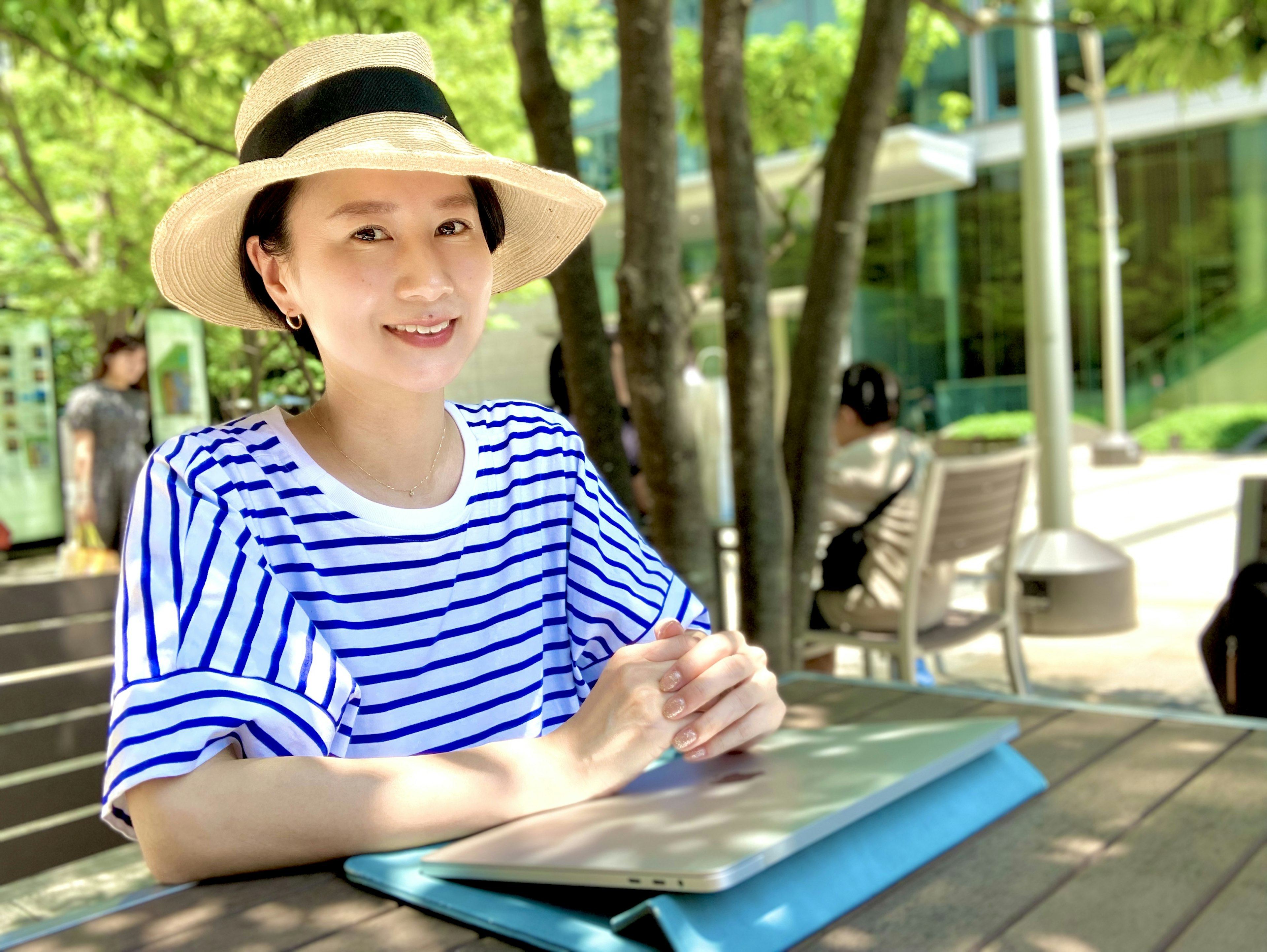 Une femme souriante assise à l'extérieur devant un ordinateur portable portant une chemise rayée et un chapeau de soleil