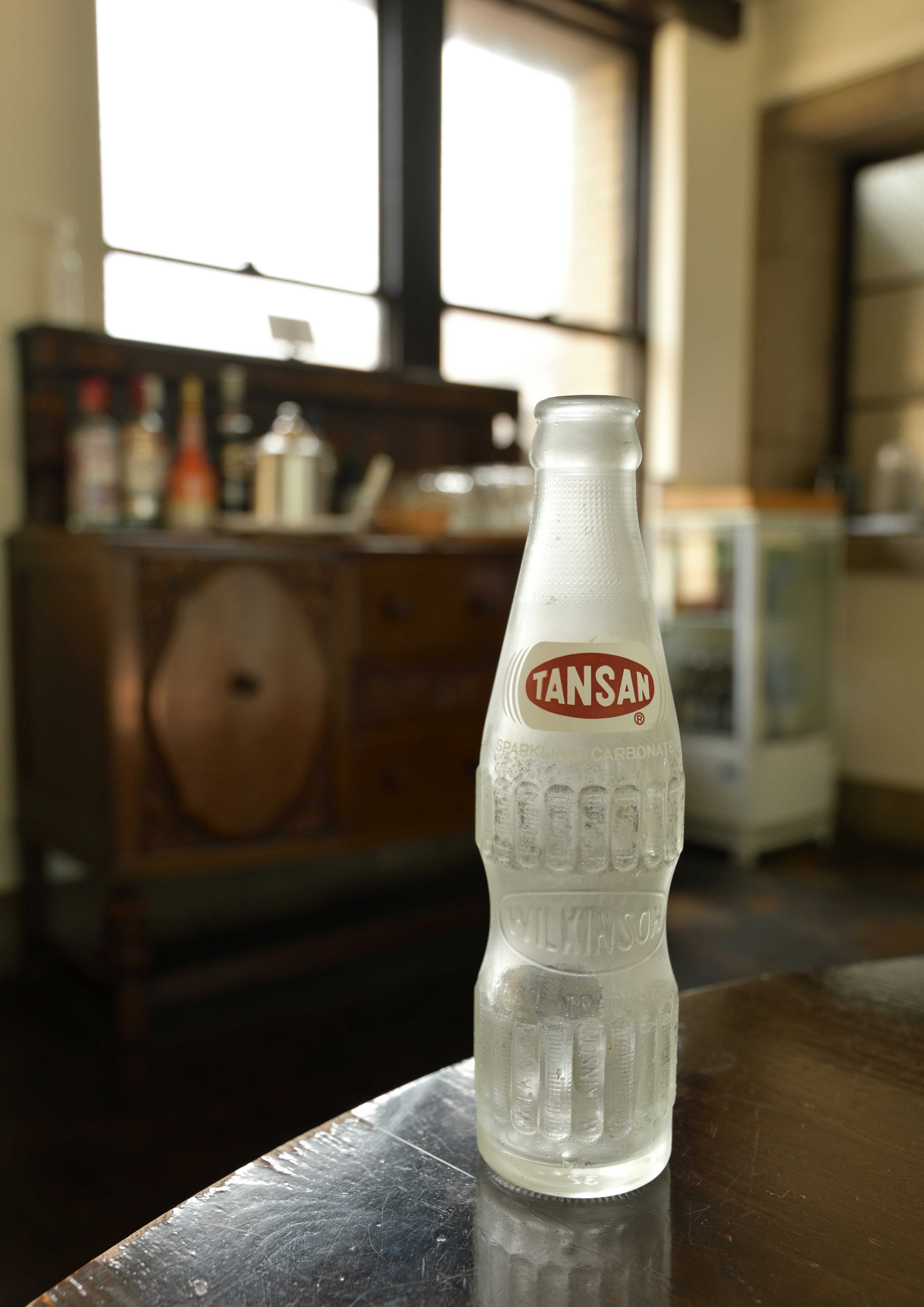 A clear beverage bottle sits on a table with a vintage wooden cabinet in the background