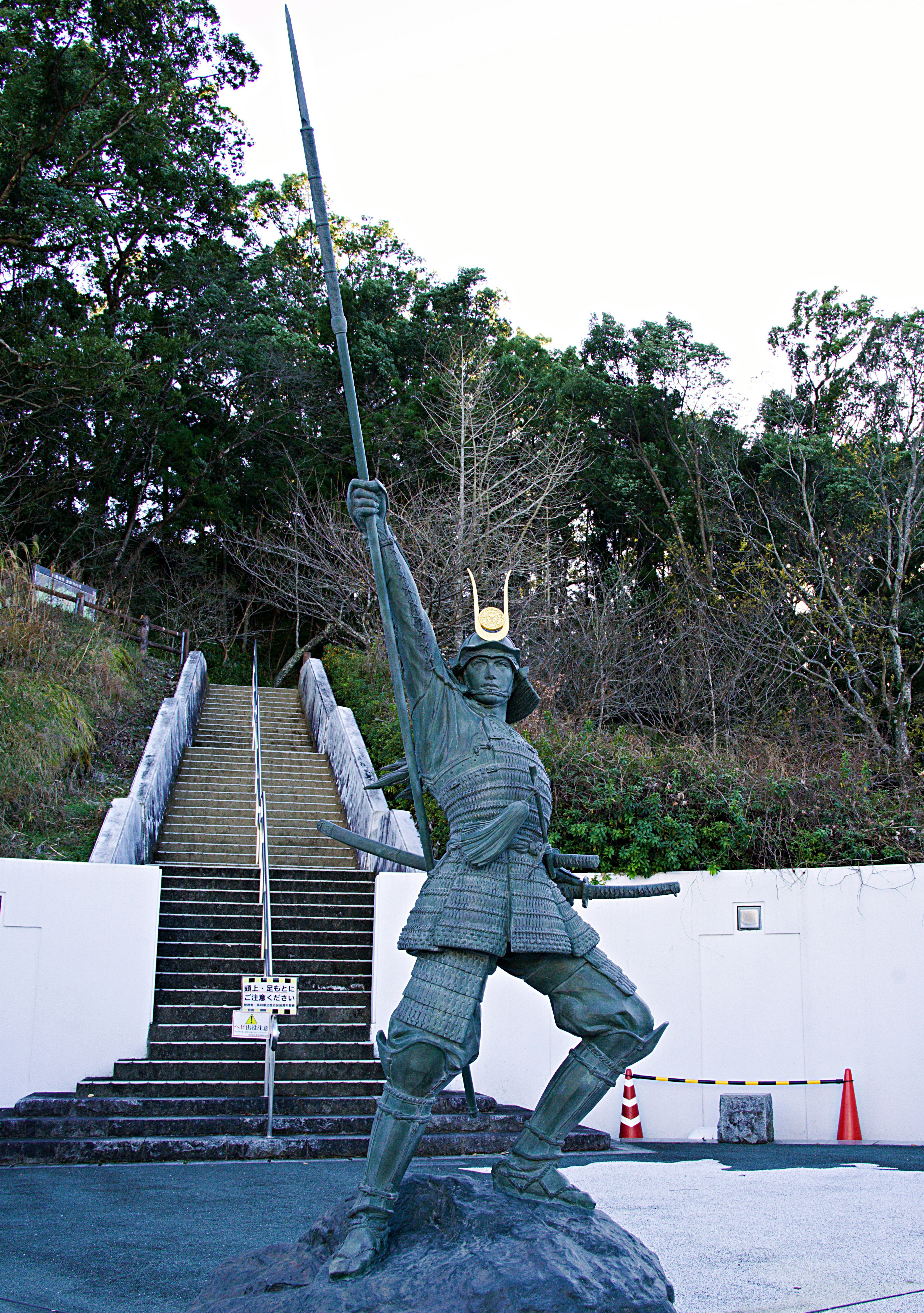 Bronze statue of a warrior holding a spear aloft near stairs and greenery