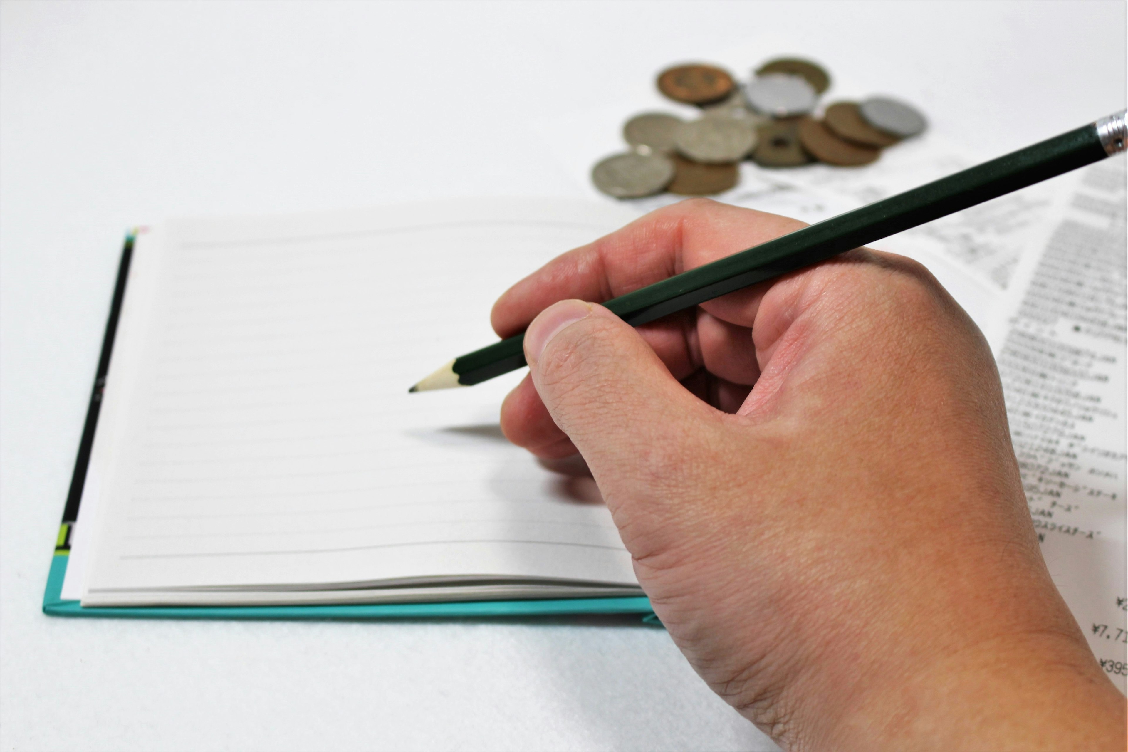 Eine Hand hält einen Stift und schreibt in ein leeres Notizbuch mit Münzen im Hintergrund