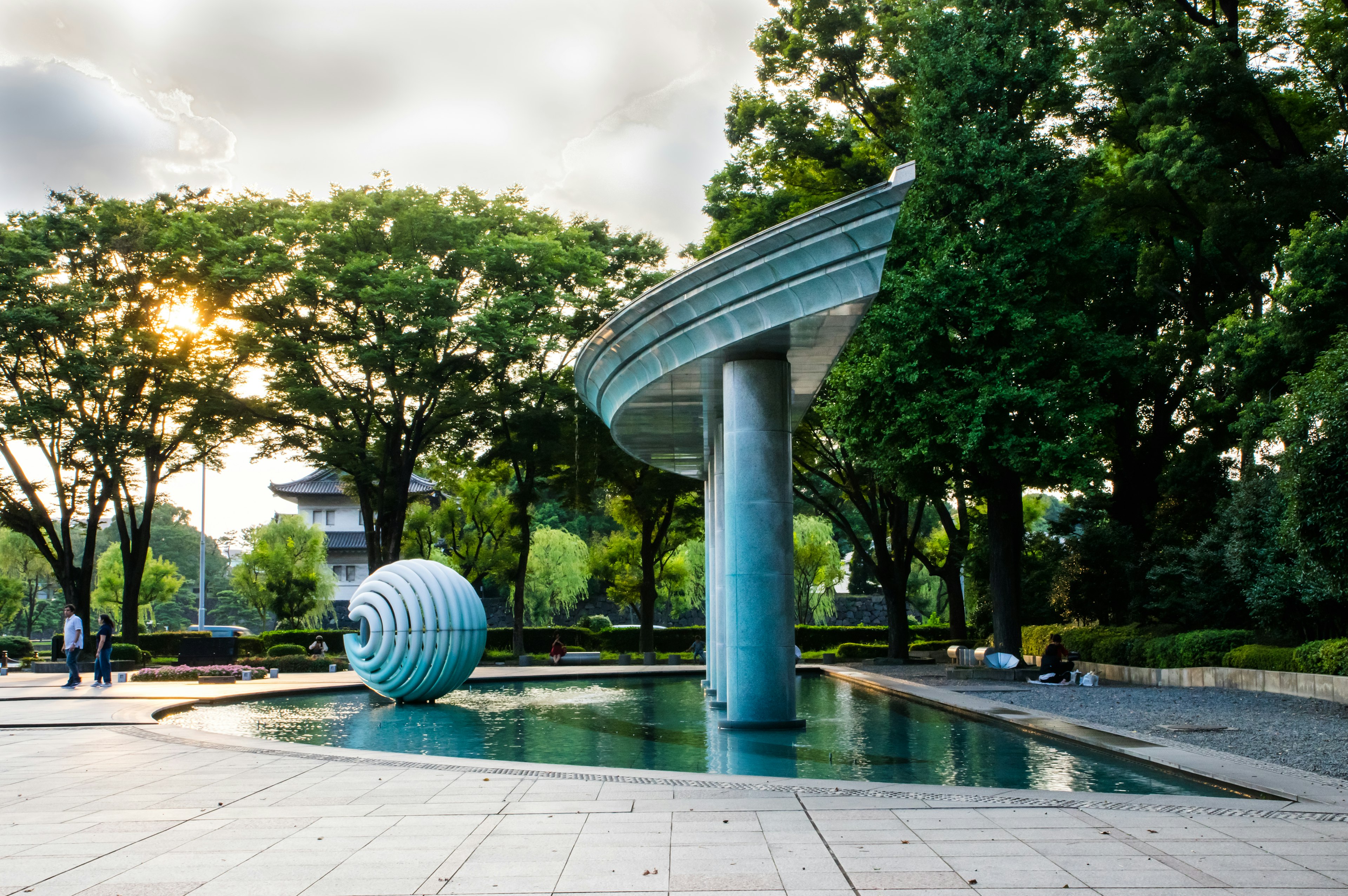 A blue circular sculpture in a park with a water feature and a curved roof